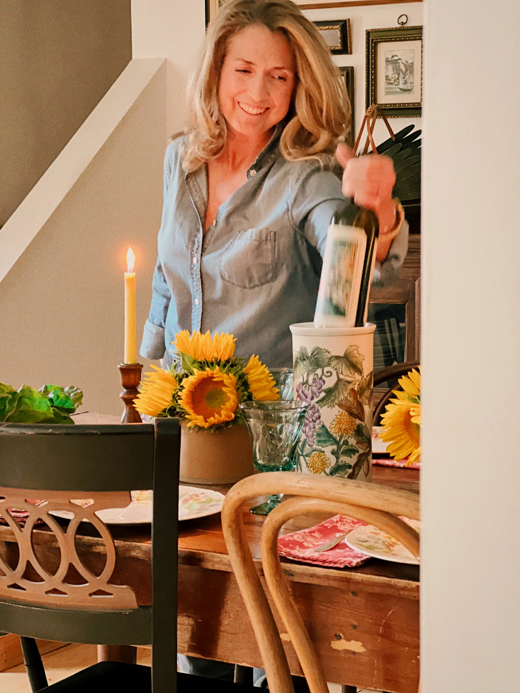 Woman setting a table with sunflowers and candles, holding a bottle of wine.