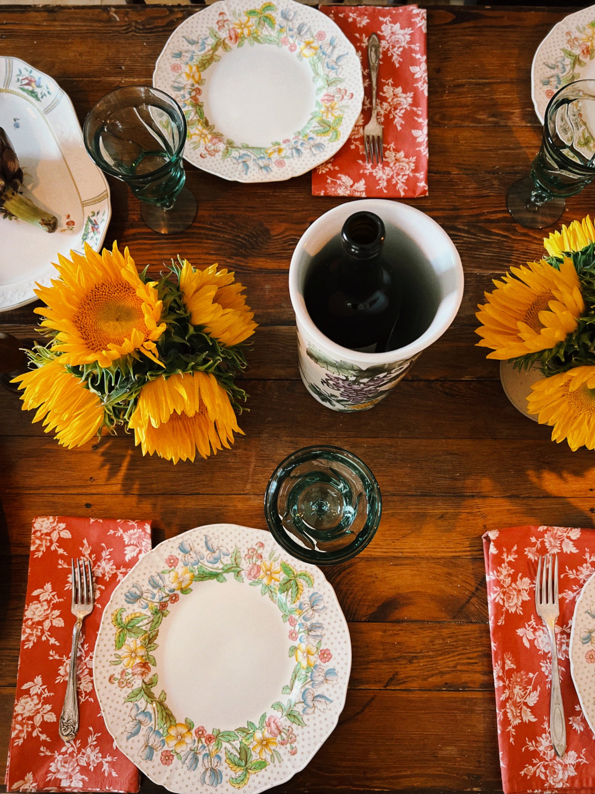 Set table with vintage floral plates, sunflowers, and red napkins on a wooden surface.