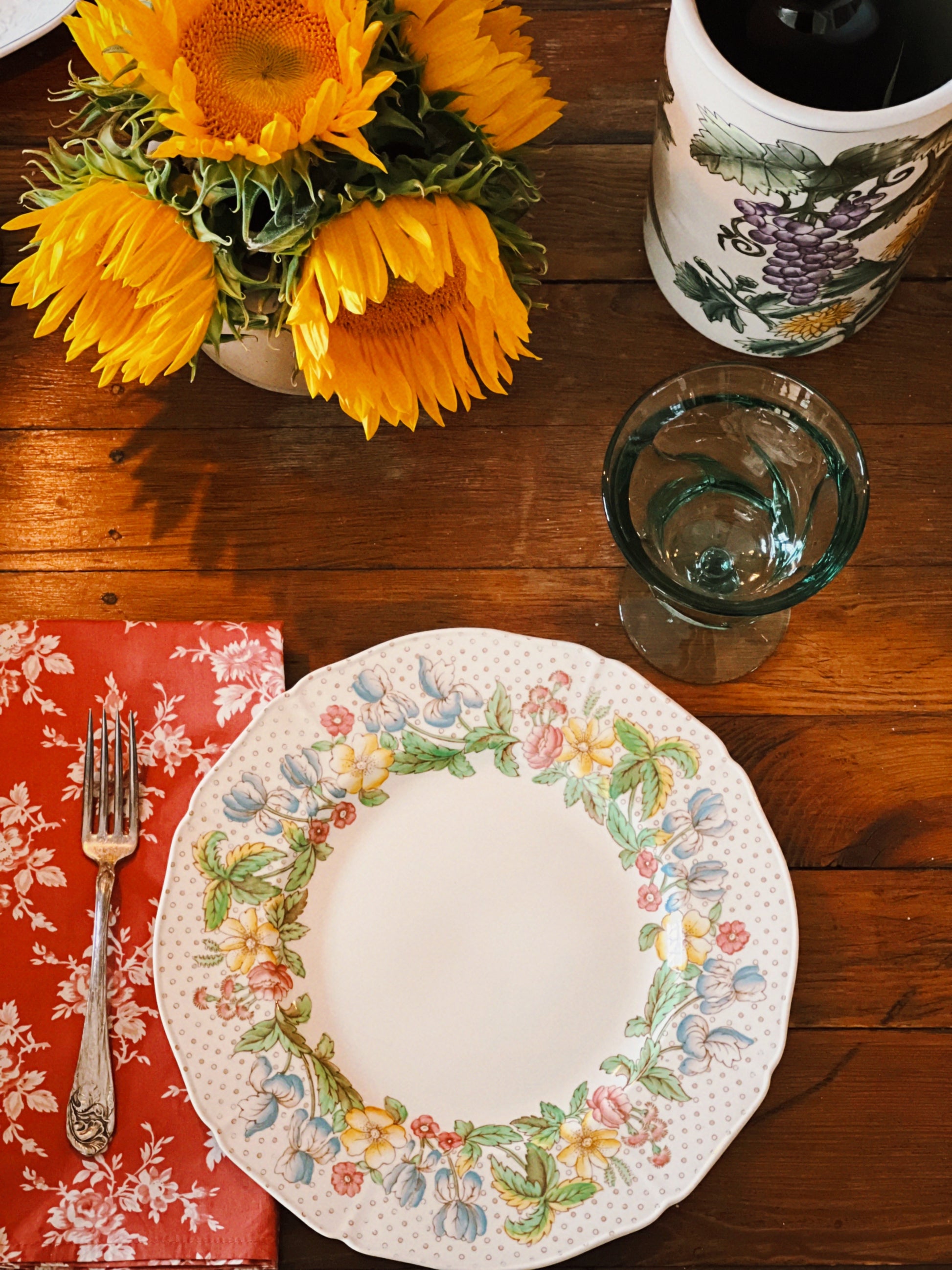 Dining table setting with vintage floral dinner plates, sunflowers, glass, and red napkin. 