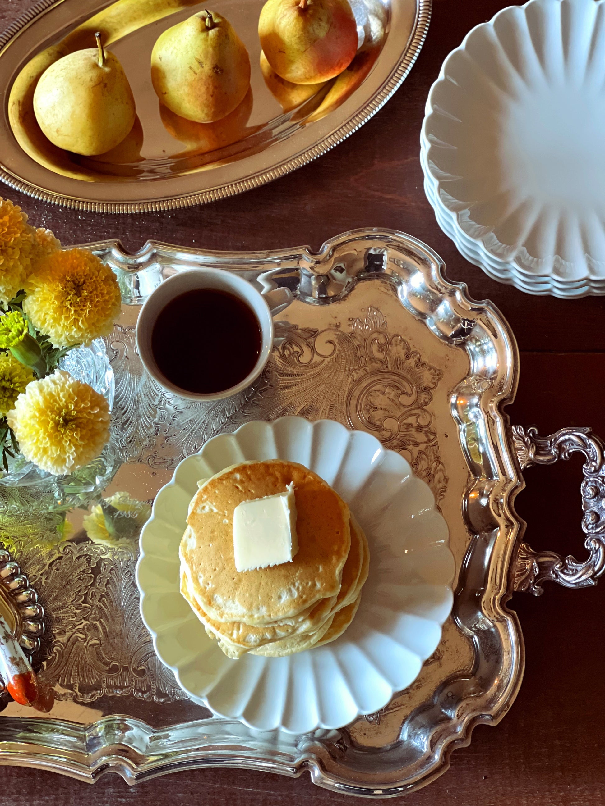 Stack of pancakes with butter on a decorative tray with a cup of coffee and pears in the background.