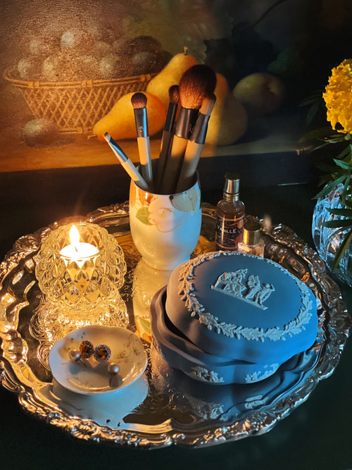 Wedgwood trinket box on a silver platter with makeup brushes, a bottle, and a candle in the background.