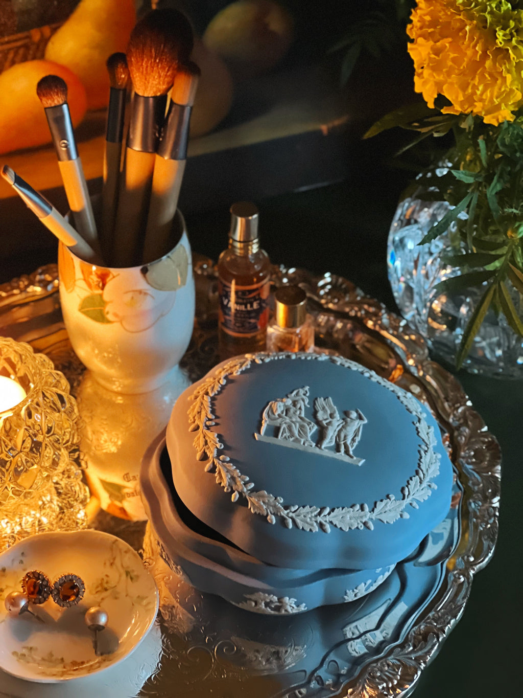 Blue Wedgwood box with intricate design on a reflective surface, surrounded by makeup brushes, perfume bottle, and flowers.