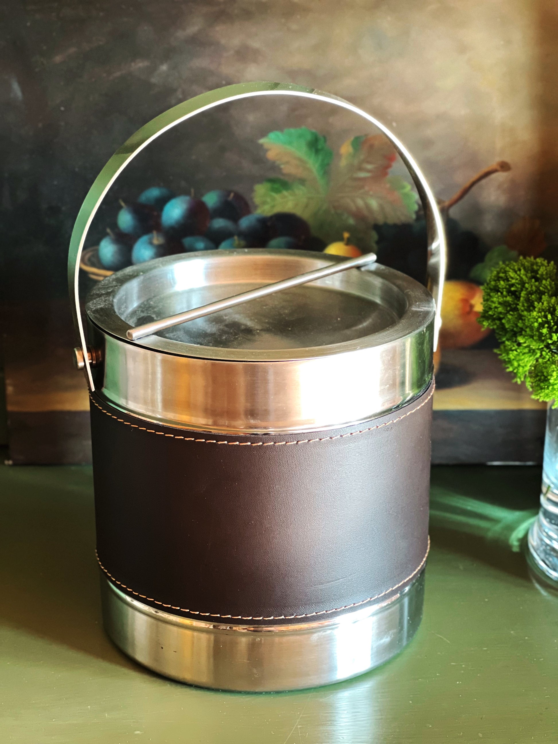 Vintage silver ice bucket with stitched faux leather wrap and bale handle displayed on a tabletop with fruit in the background.