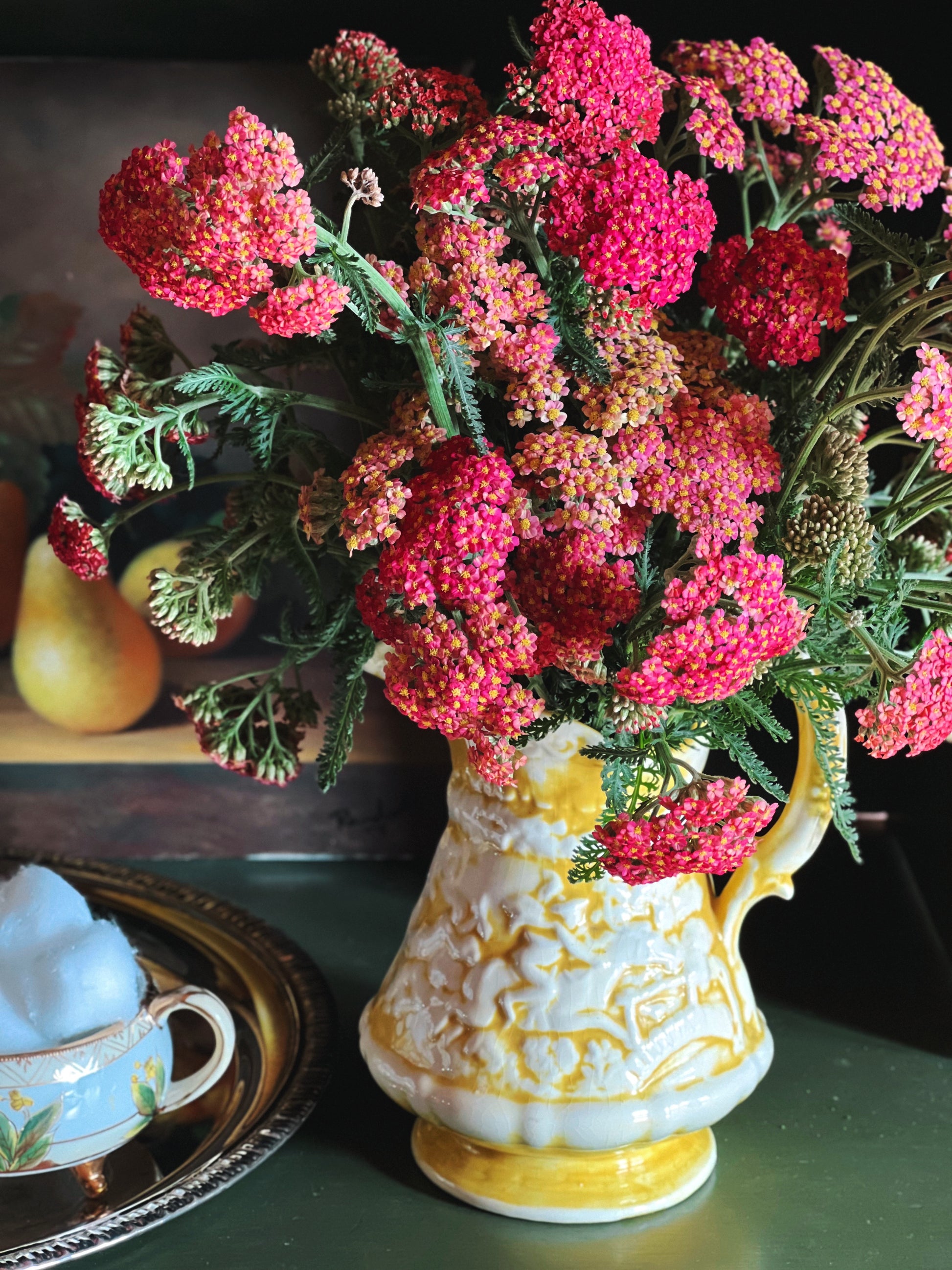 Handmade vintage yellow ceramic pitcher filled with pink yarrow flowers beside antique treasures and citron pears