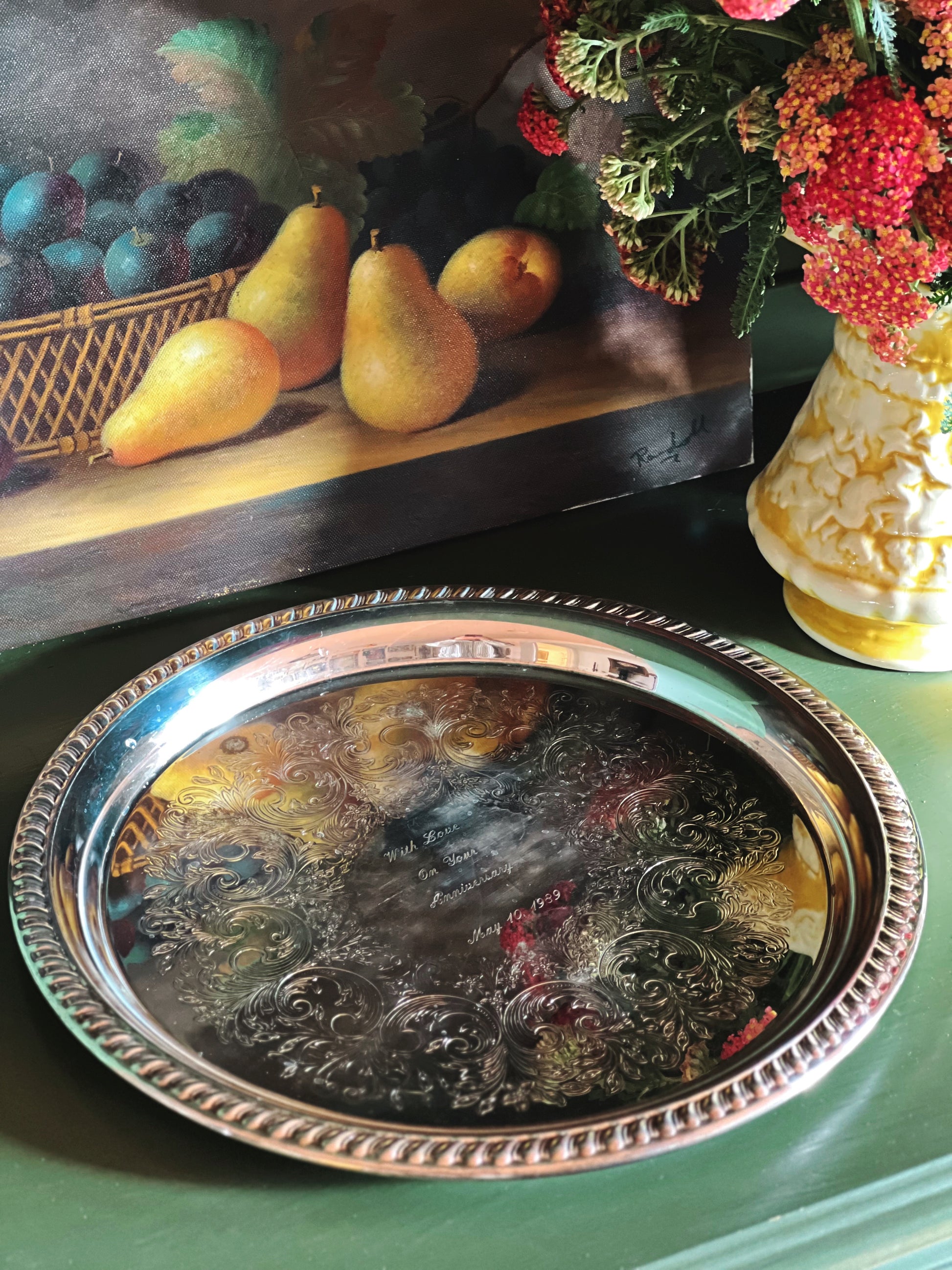 Decorative silver tray on a table with a painting of pears and a vase of flowers in the background.