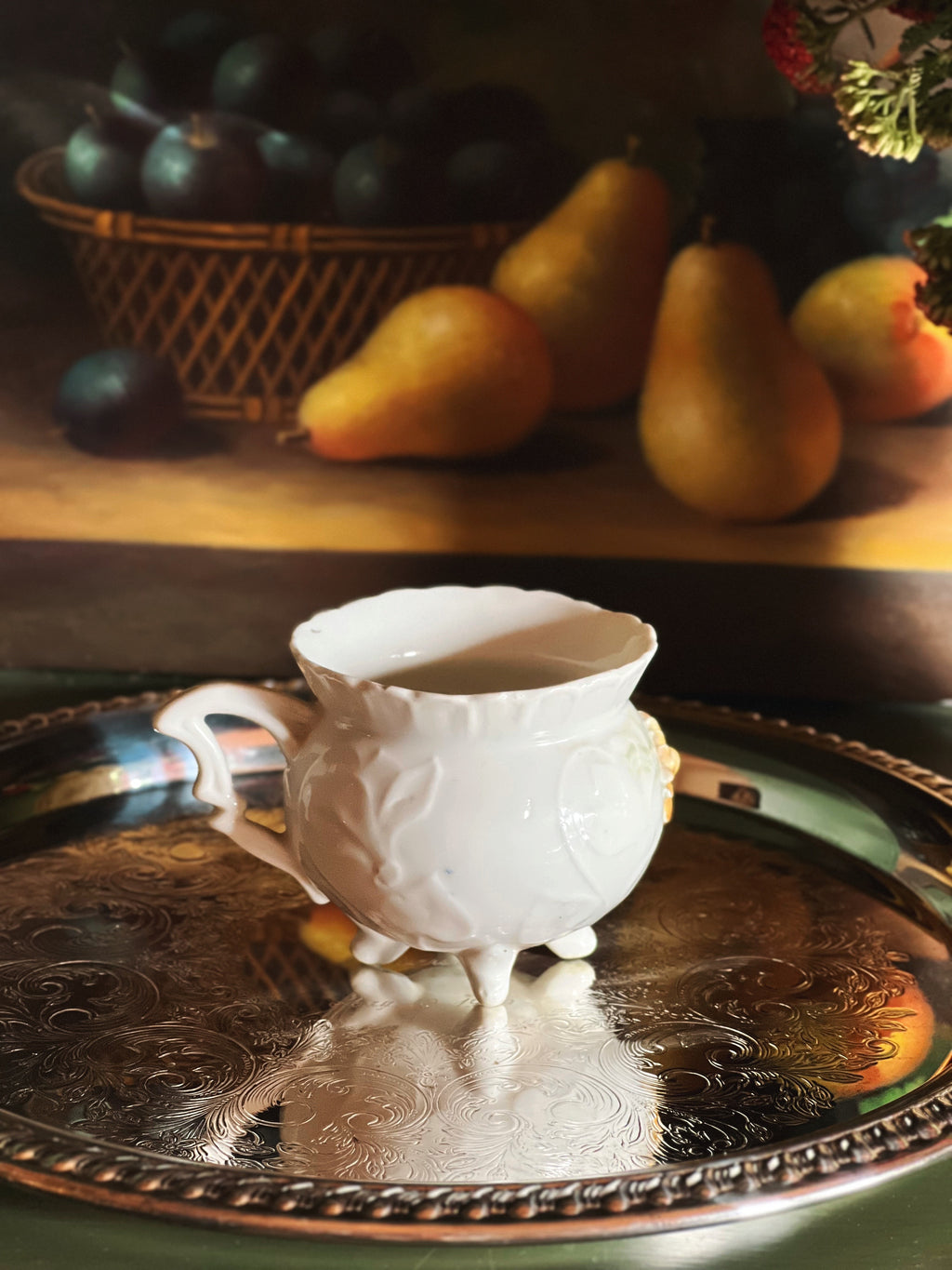 White decorative cup on a silver tray with pears and plums in the background