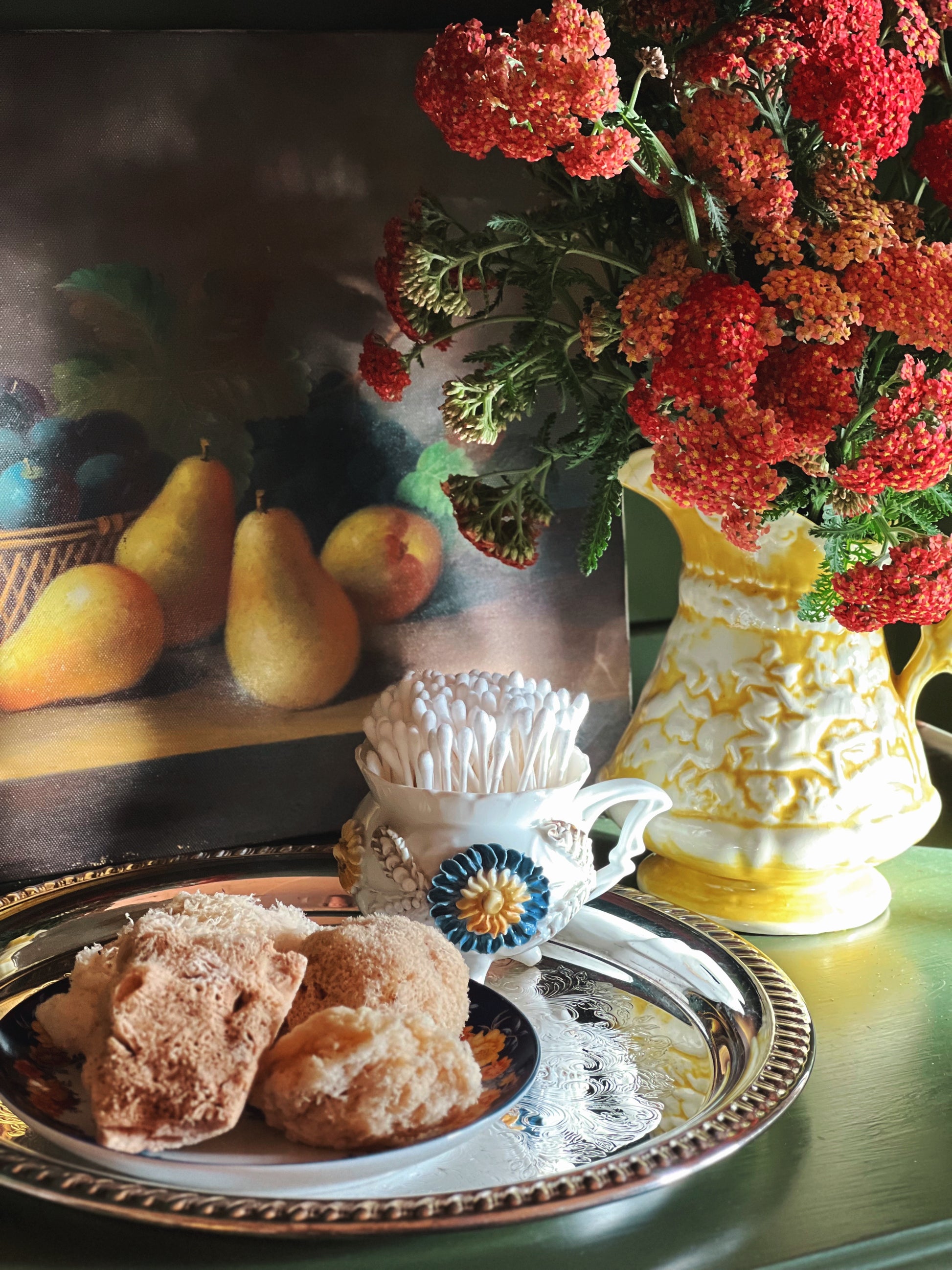 Handmade vintage yellow ceramic pitcher filled with pink yarrow flowers beside antique treasures and citron pears