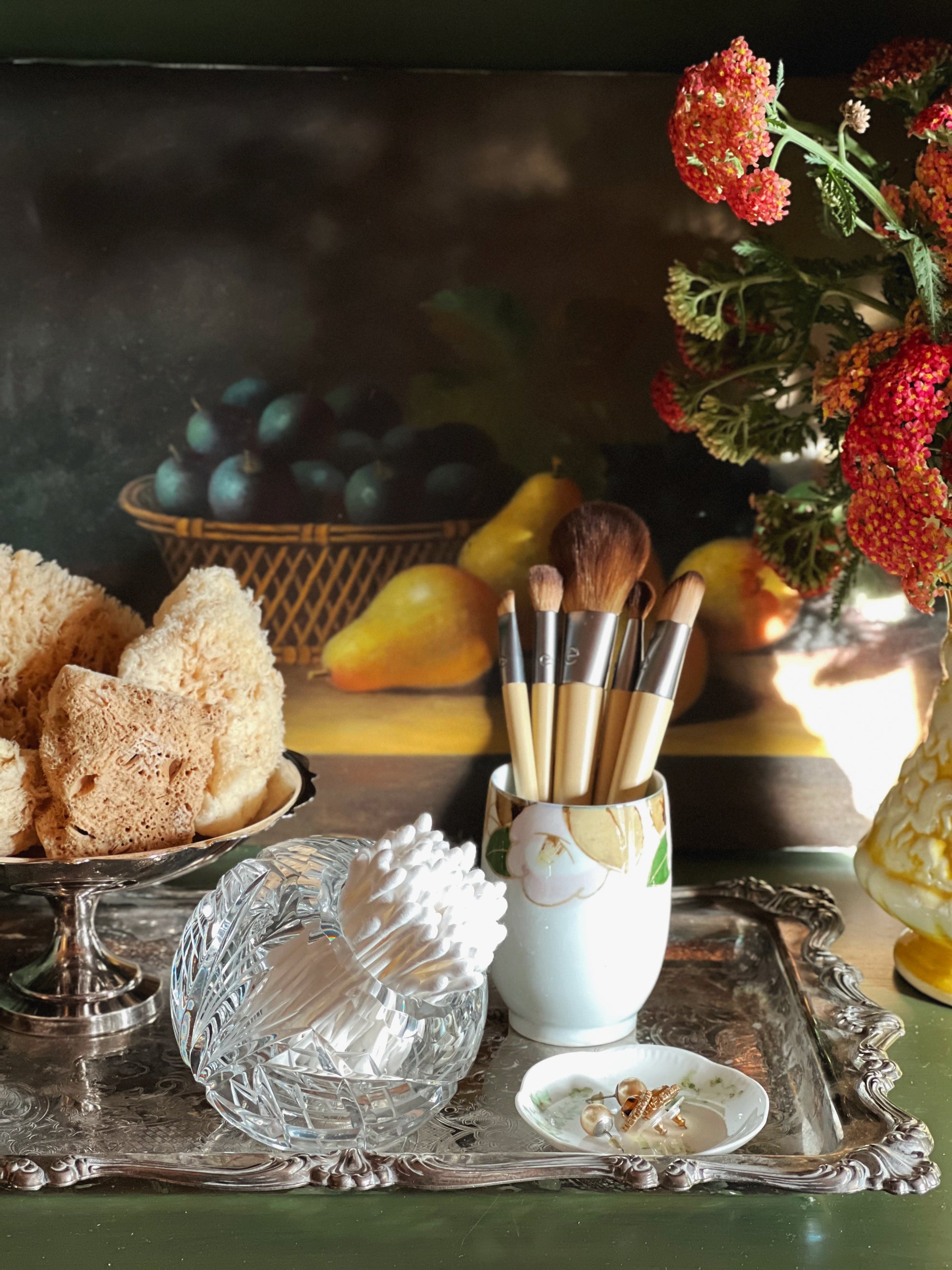 Makeup brushes in a holder on a decorative tray with fruit and flowers in the background