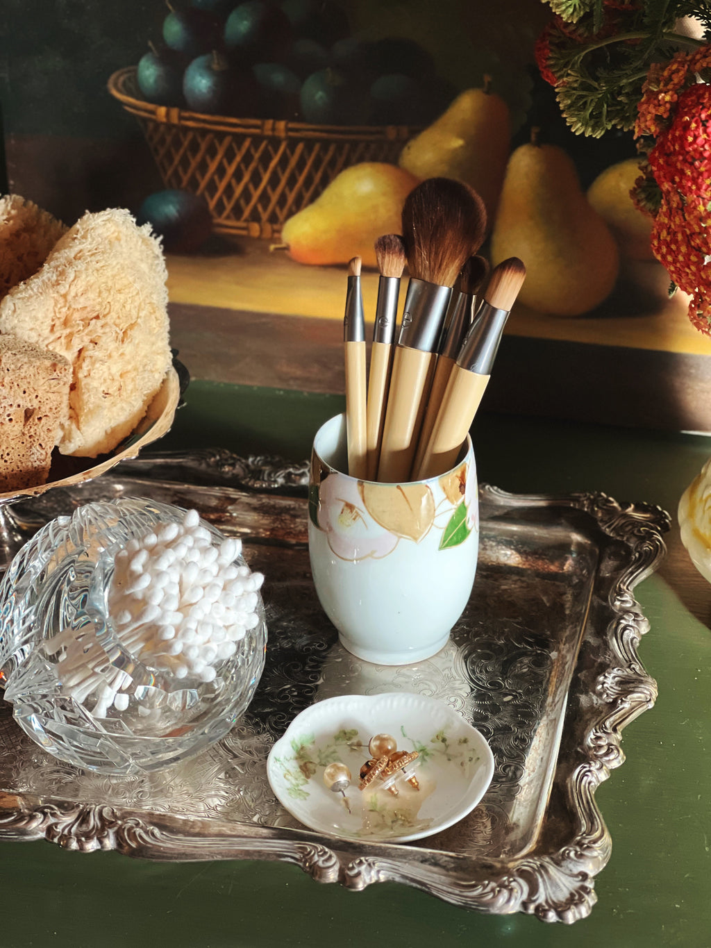 Set of makeup brushes in a decorative cup on a ornate tray with fruits and bread in the background