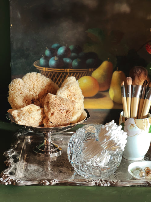 Vintage setting with sponges, a crystal dish, and makeup brushes on a reflective surface.