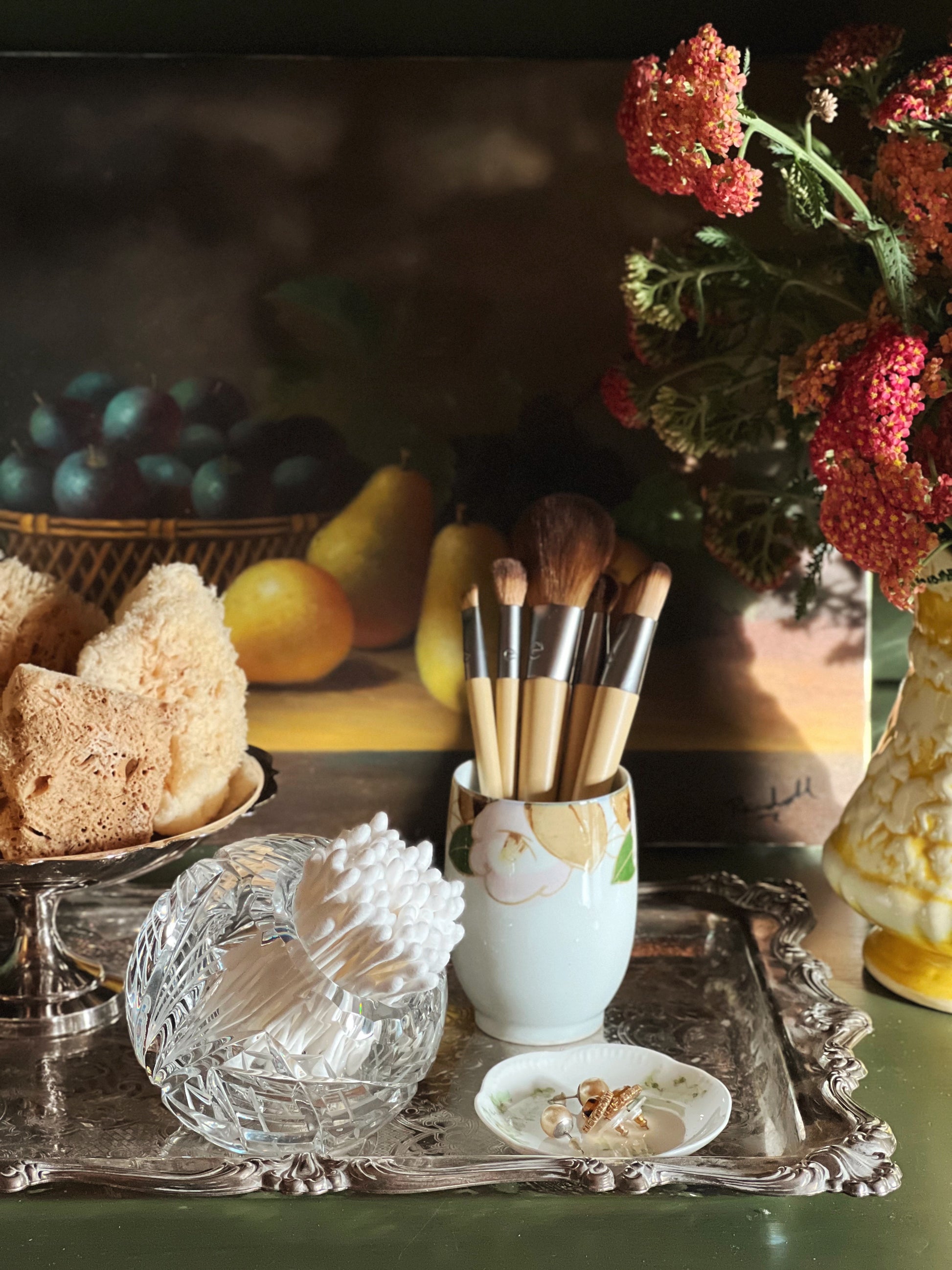 Makeup brushes in a holder on a decorative tray with fruit and flowers in the background