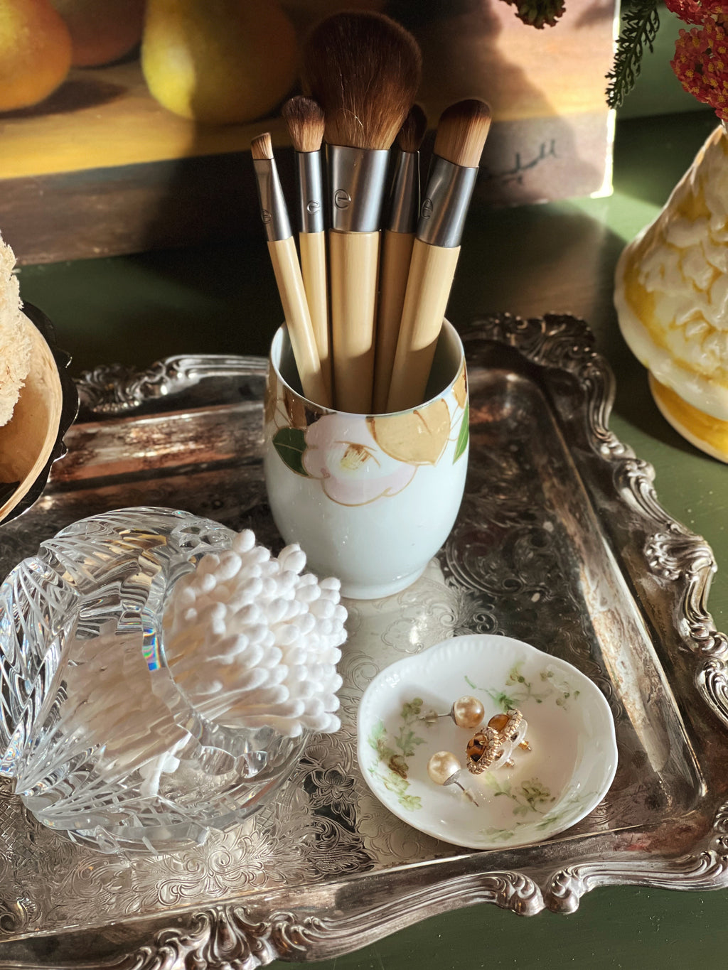 Makeup brushes in a decorative cup on a silver tray with fruit and a vase in the background
