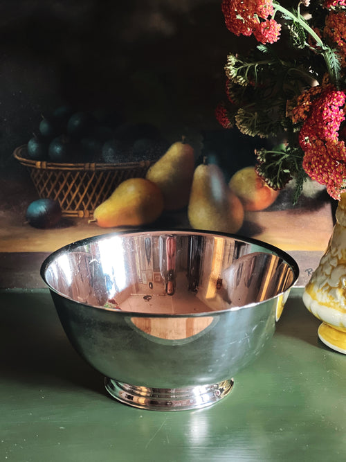Silver bowl on a wooden surface with pears and a vase of flowers in the background.