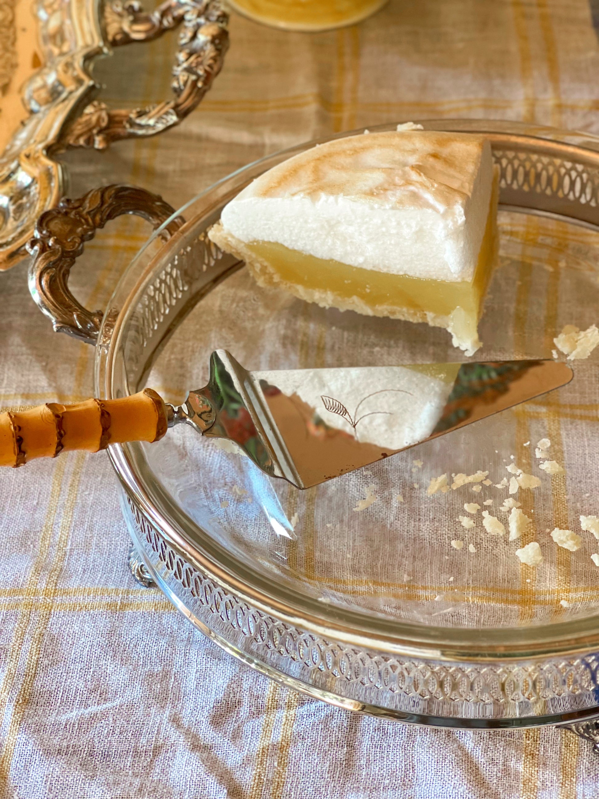 Slice of pie in a Fire King glass pie plate on a silver casserole server with a decorative handle, on a patterned tablecloth.