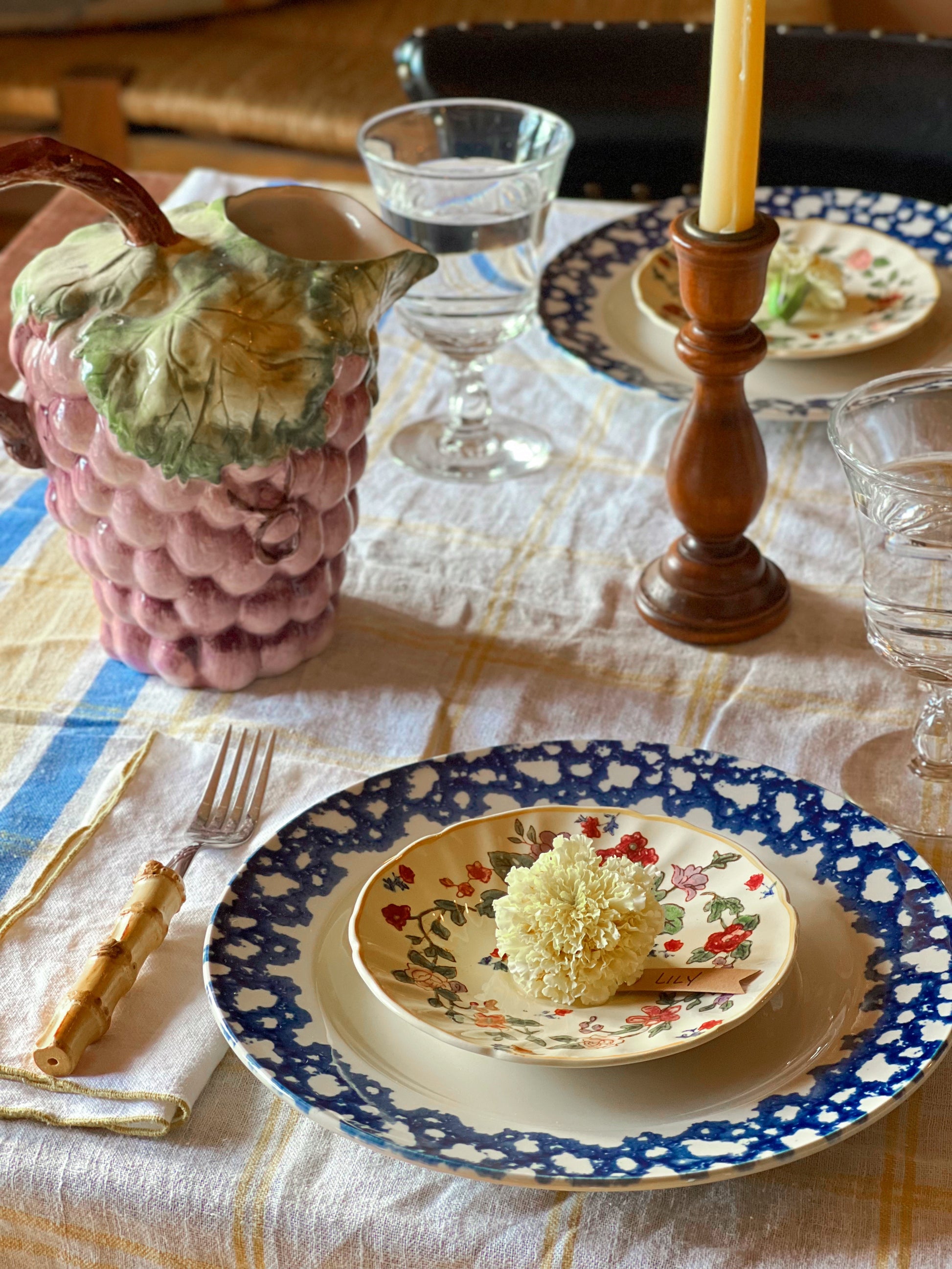 Dining table setting with floral plates, a pink pineapple-shaped pitcher, and a wooden candlestick.