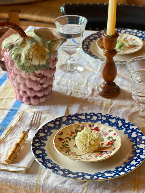 Dining table setting with floral plates, a pink pineapple-shaped pitcher, and a wooden candlestick.