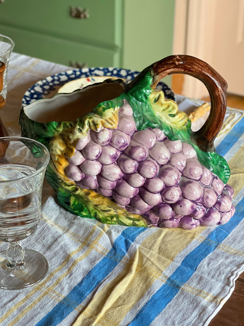 Decorative ceramic pitcher shaped like a bunch of grapes on a table with glasses and a plate.