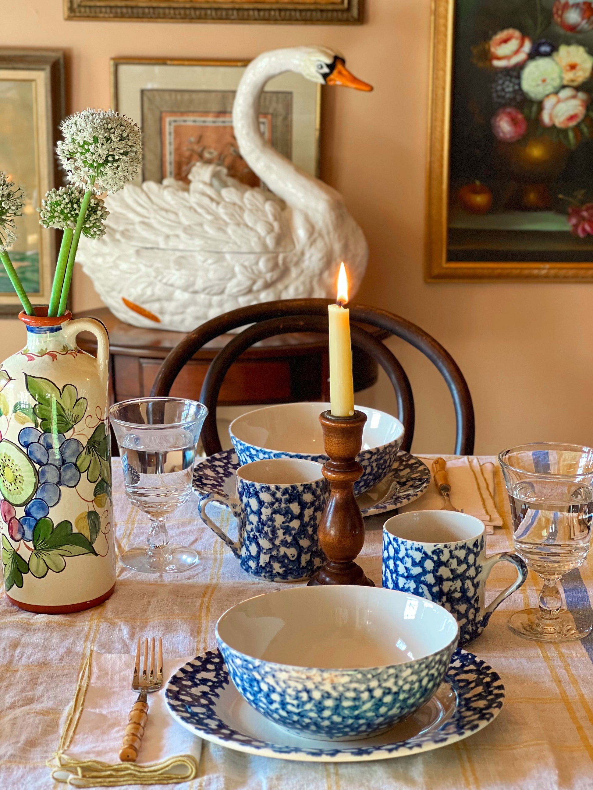 Dining table setting with swan-shaped tureen, floral vase, and candle in a room with framed pictures on the wall.