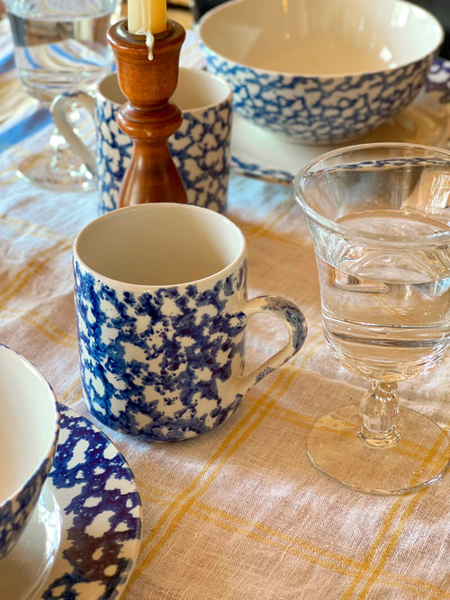Table setting with blue and white ceramic mug, glass, and candle on a checkered tablecloth.