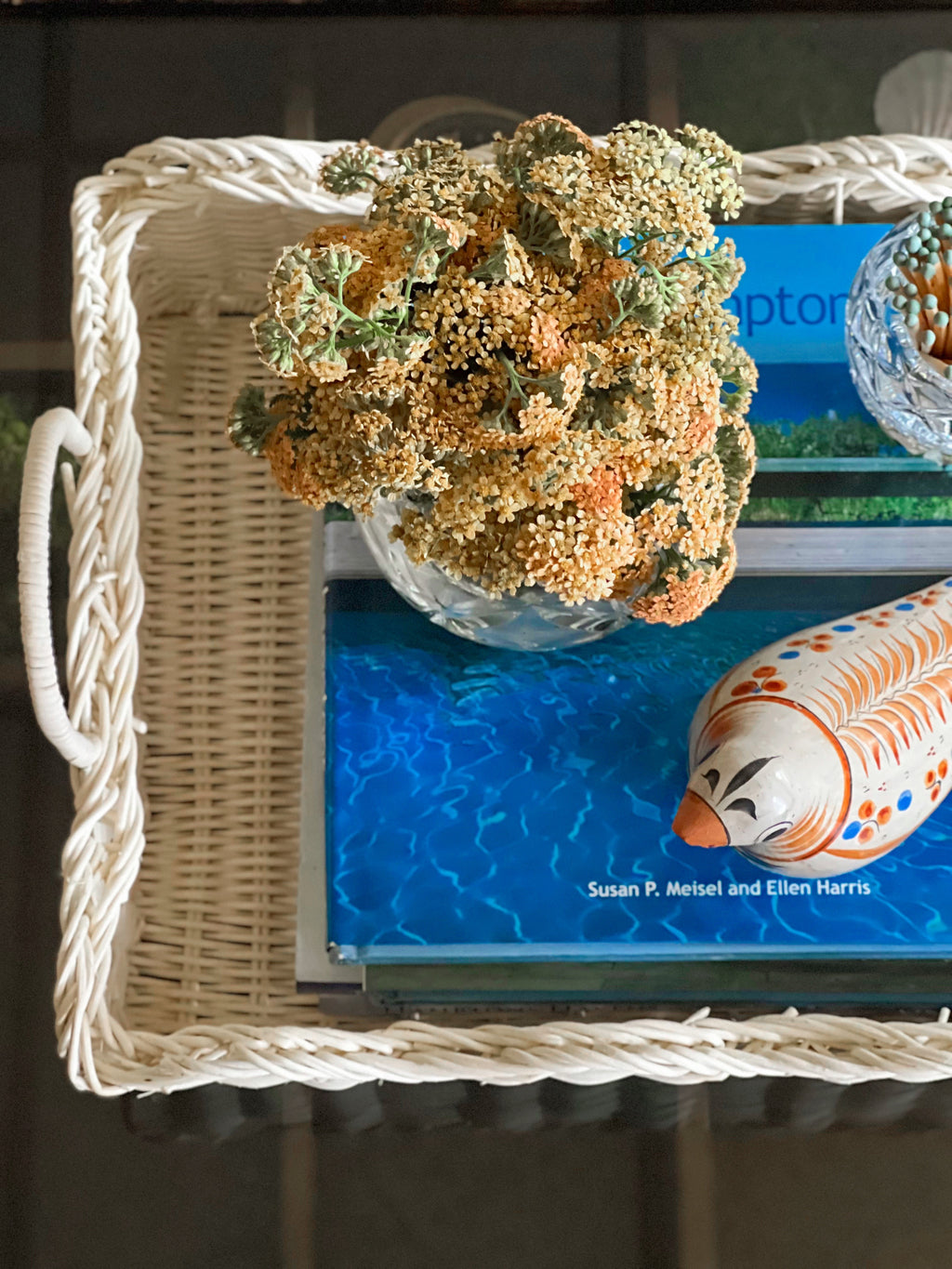 Overhead view of a white wicker tray on a coffee table with coastal books and decorative elements