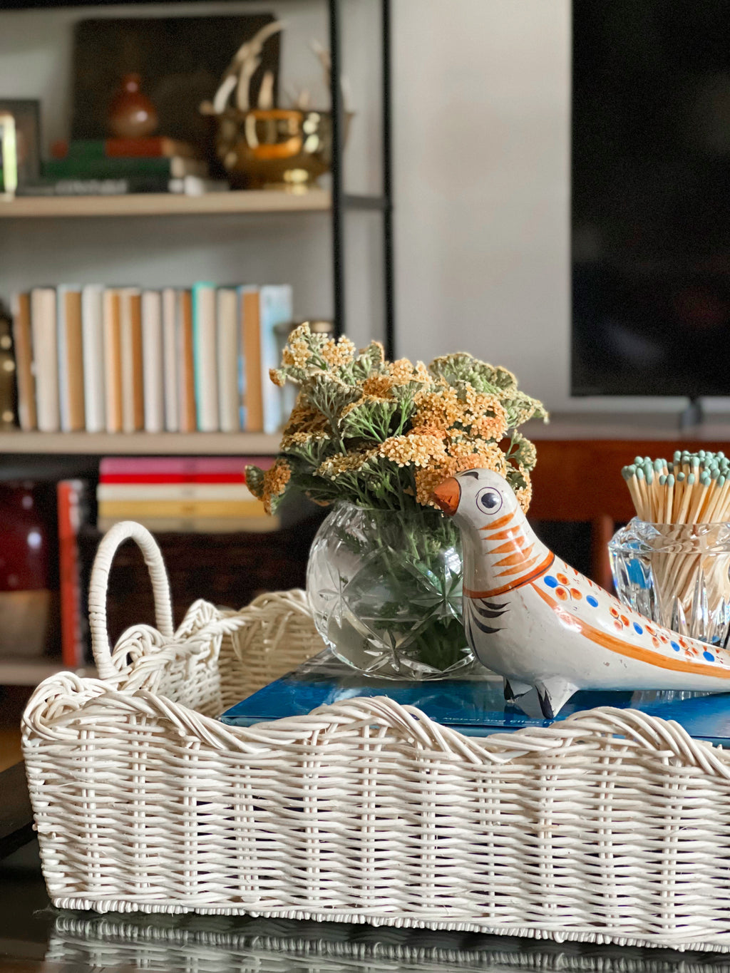Decorative setting with a wicker basket, ceramic bird, vase with flowers, and books in a room.