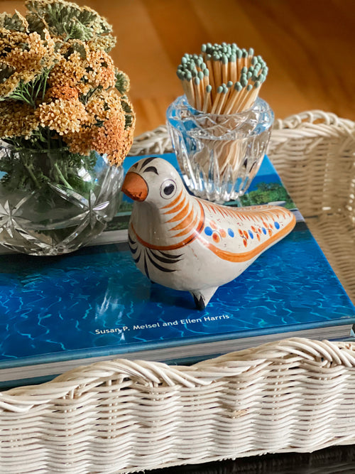 Decorative  Mexican Tonal bird figurine on a book in a white wicker tray with a vase of flowers and matches in the background.
