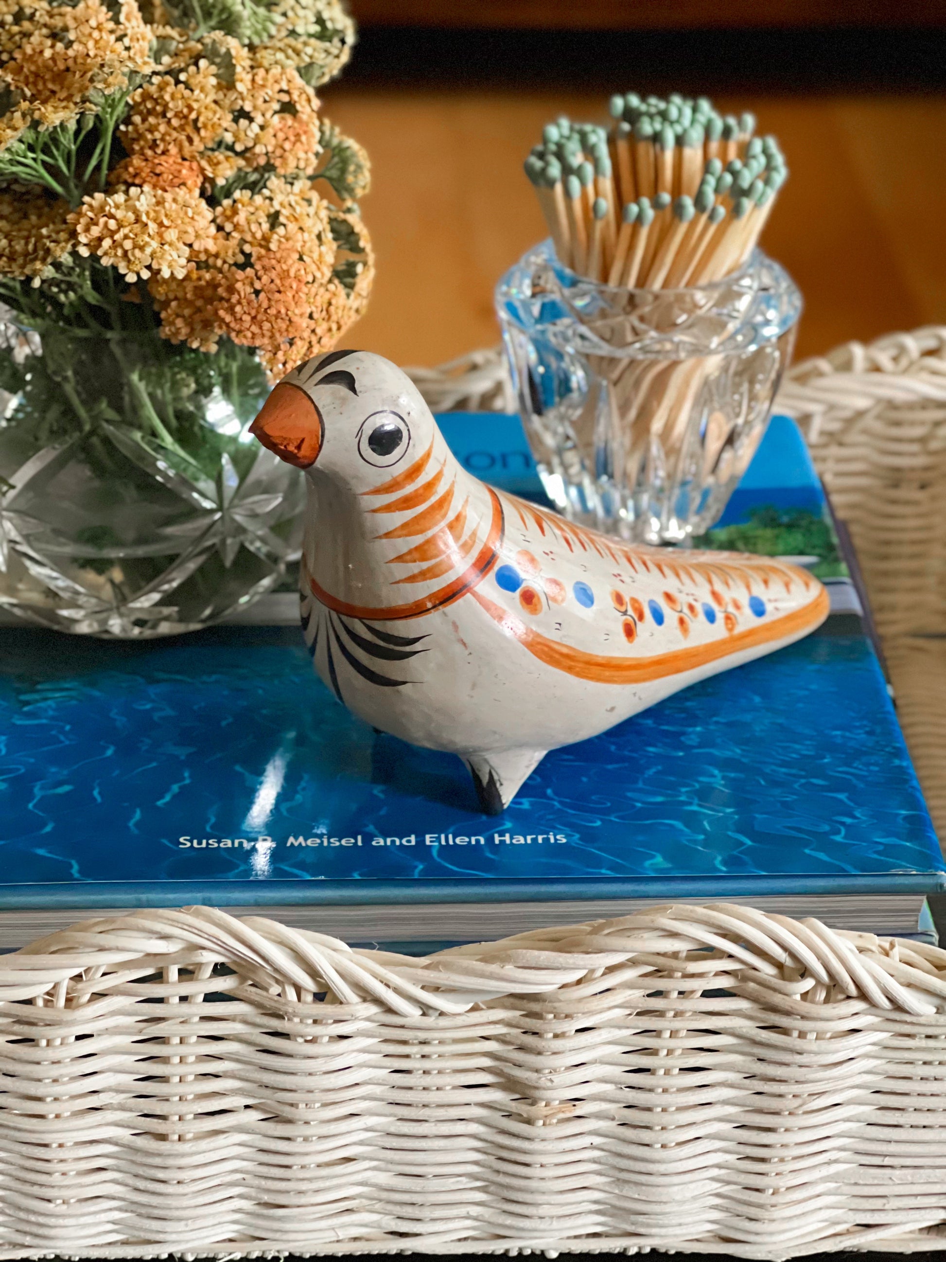 Decorative Tonal Mexican bird figurine on a book with a vase of flowers and matchstick holder in the background.