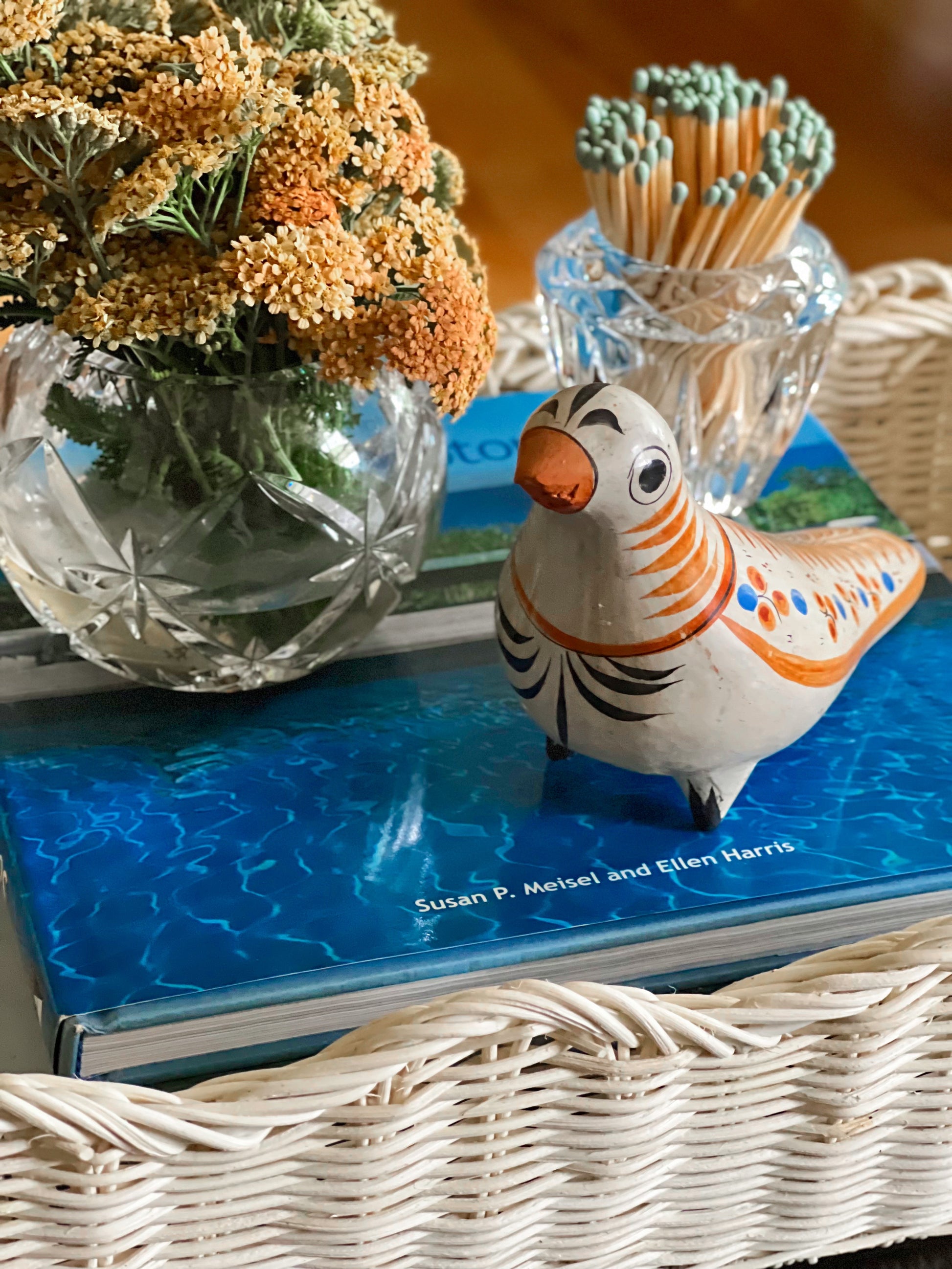 Decorative scene with a Tonala Mexico ceramic bird, vase of flowers, and book on a surface.