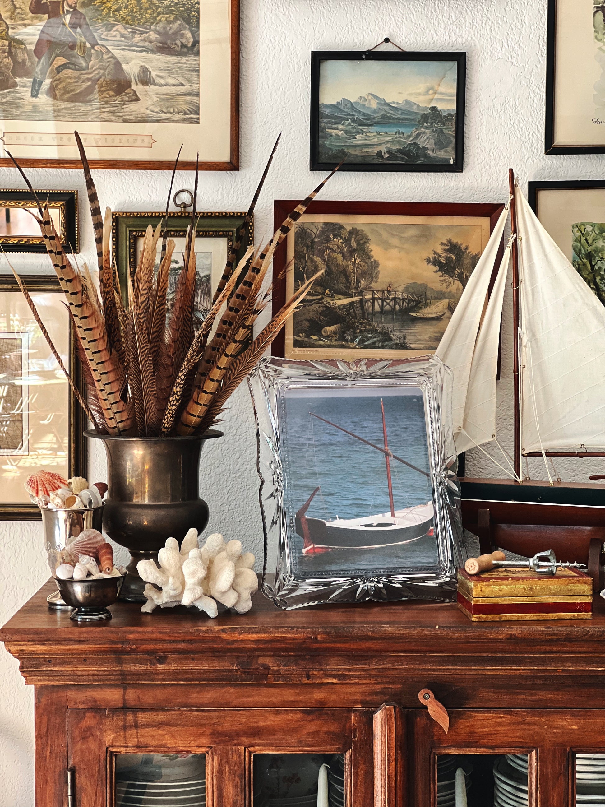 Decorative shelf with feather arrangement, framed pictures, and a model sailboat.