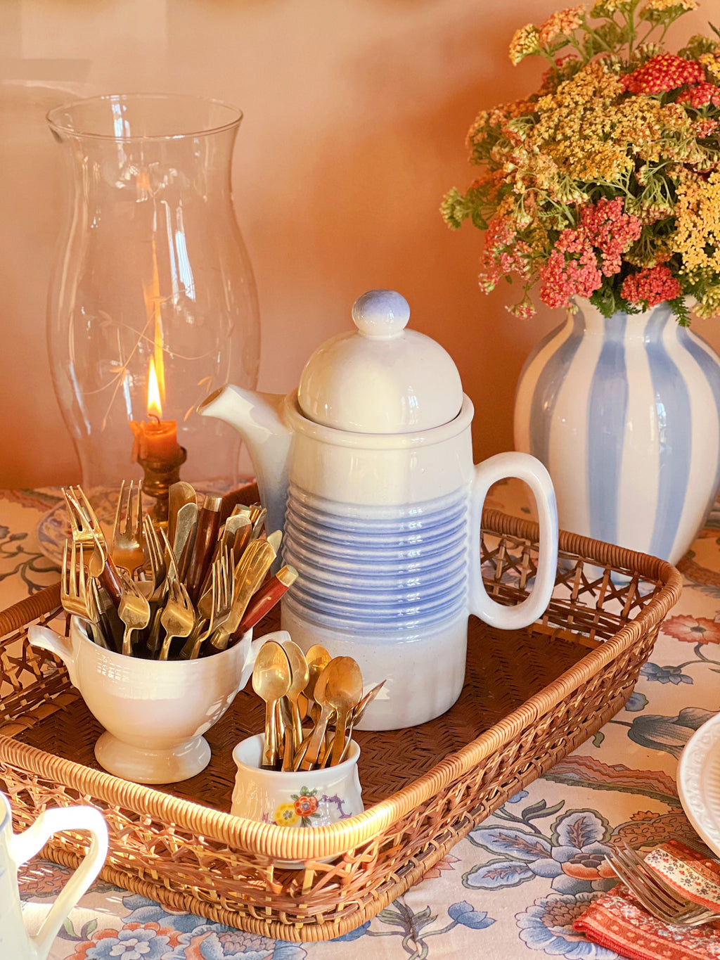 Decorative table setting with a teapot, candle, and floral arrangement on a wicker tray.