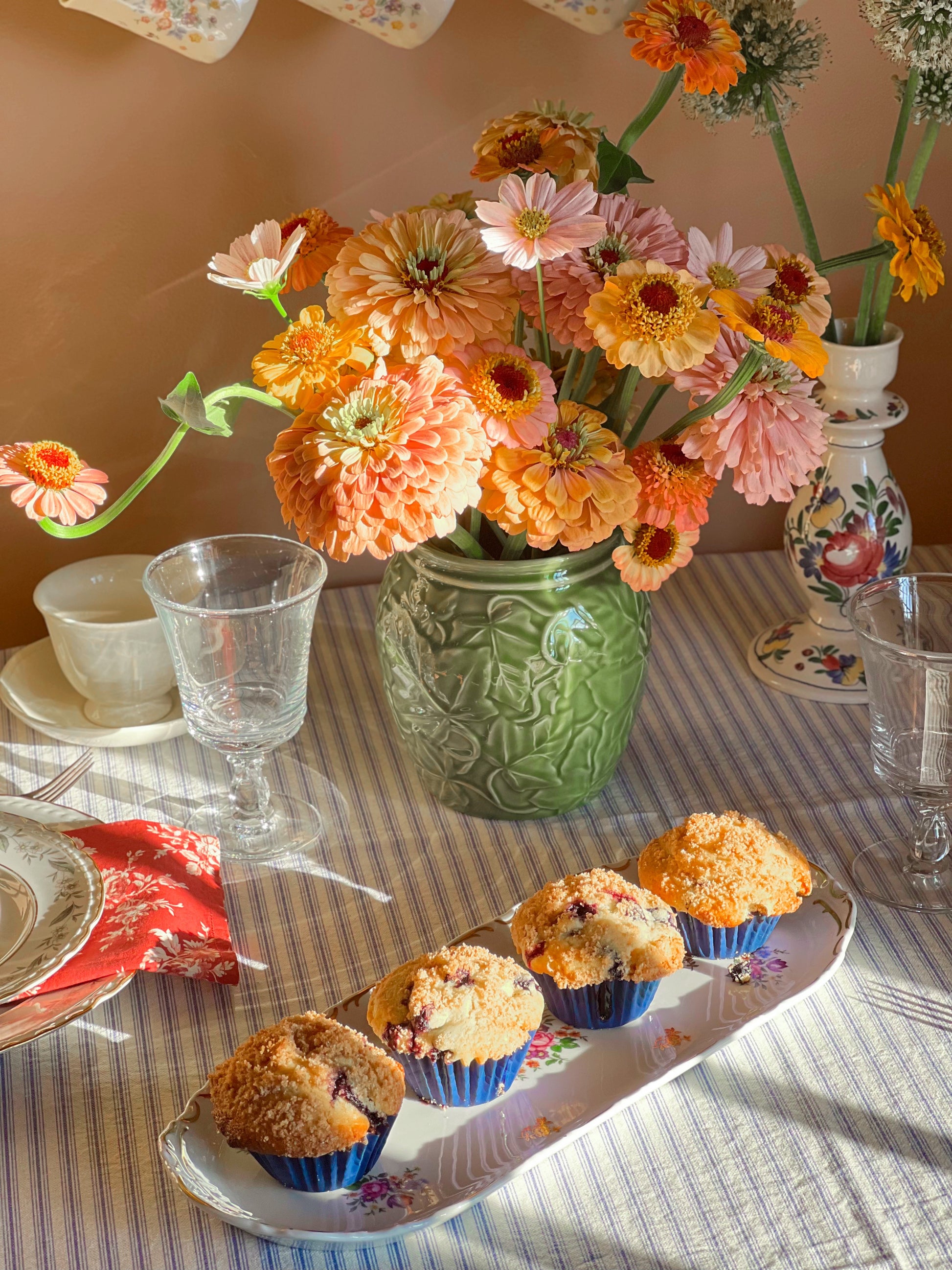 Table setting with muffins on a German porcelain tray, flowers, and glasses on a striped tablecloth.