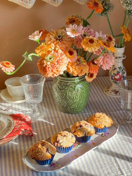 Table setting with muffins on a German porcelain tray, flowers, and glasses on a striped tablecloth.