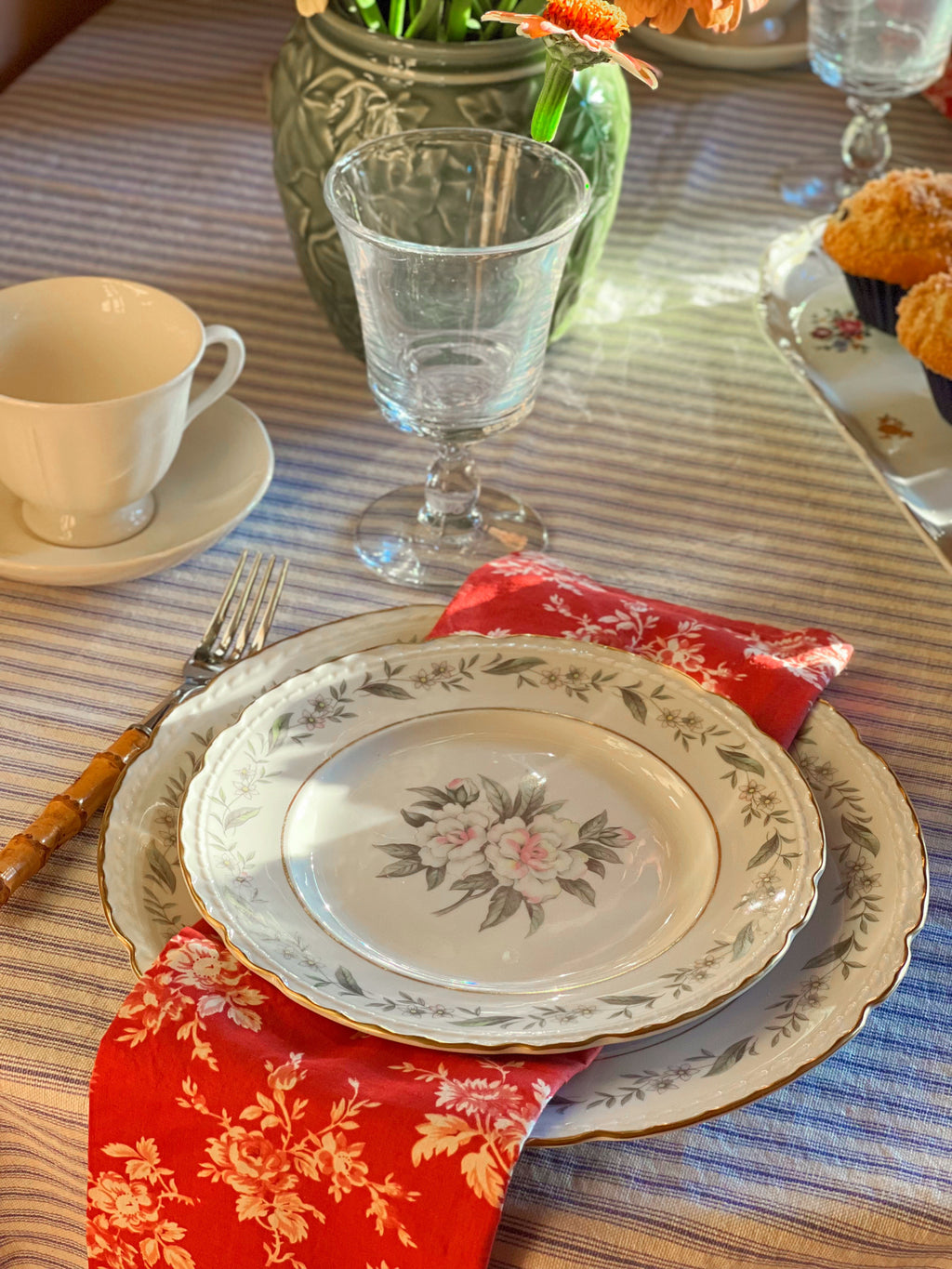 Set table with floral plates, glasses, and a cup on a striped tablecloth.