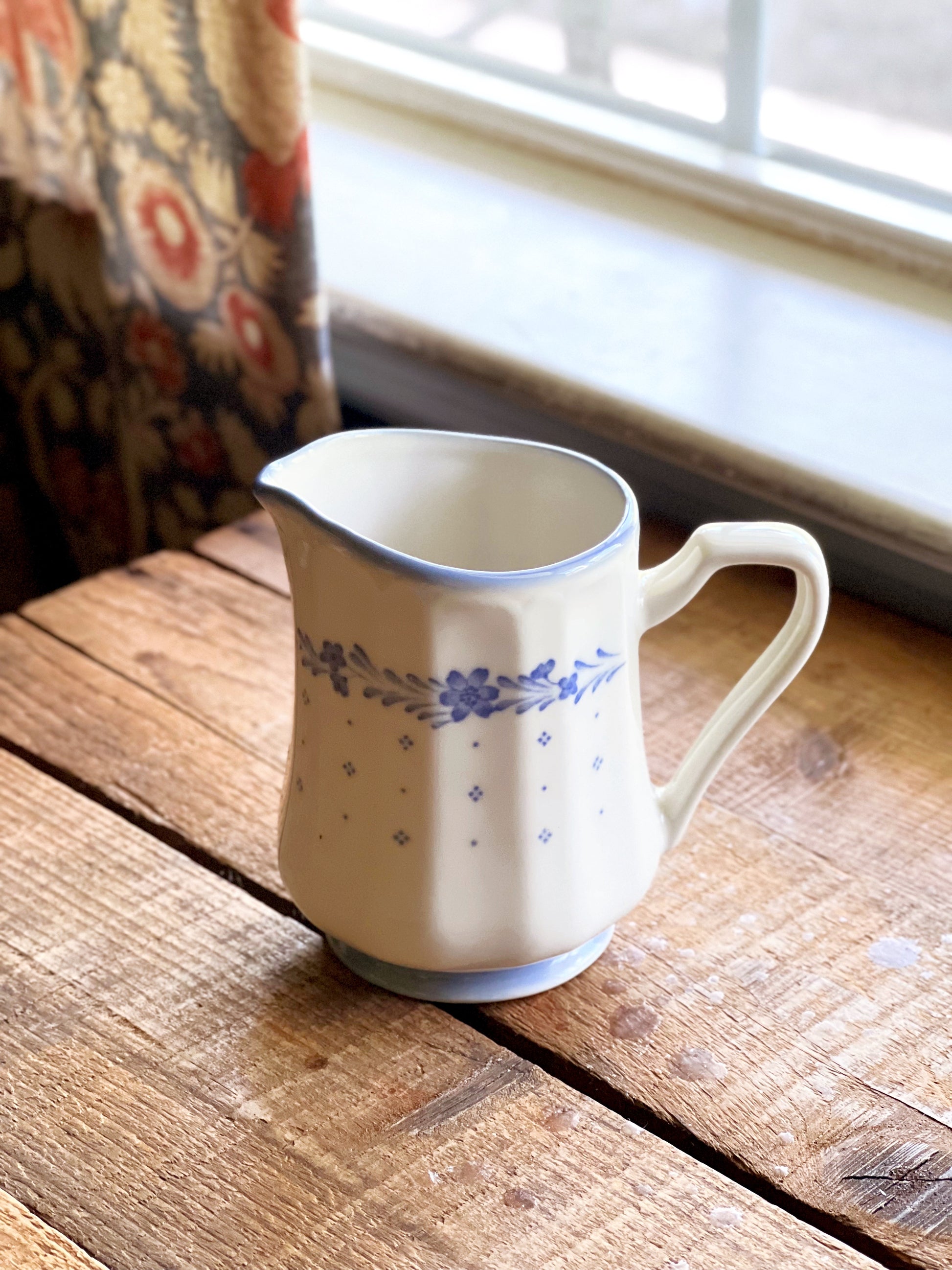White ceramic pitcher with blue floral patterns on a wooden surface near a window.