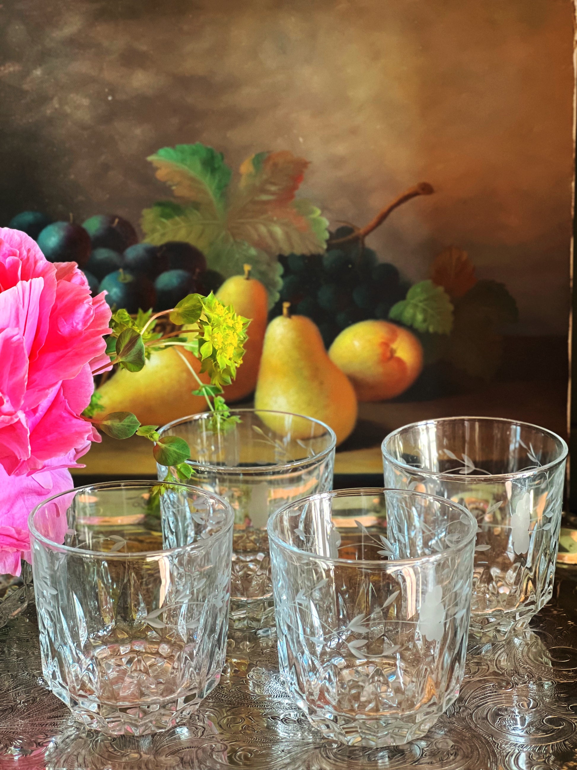 Set of four vintage etched crystal double old fashioned glasses arranged on a moody tabletop with pink peonies and fruit in the background.