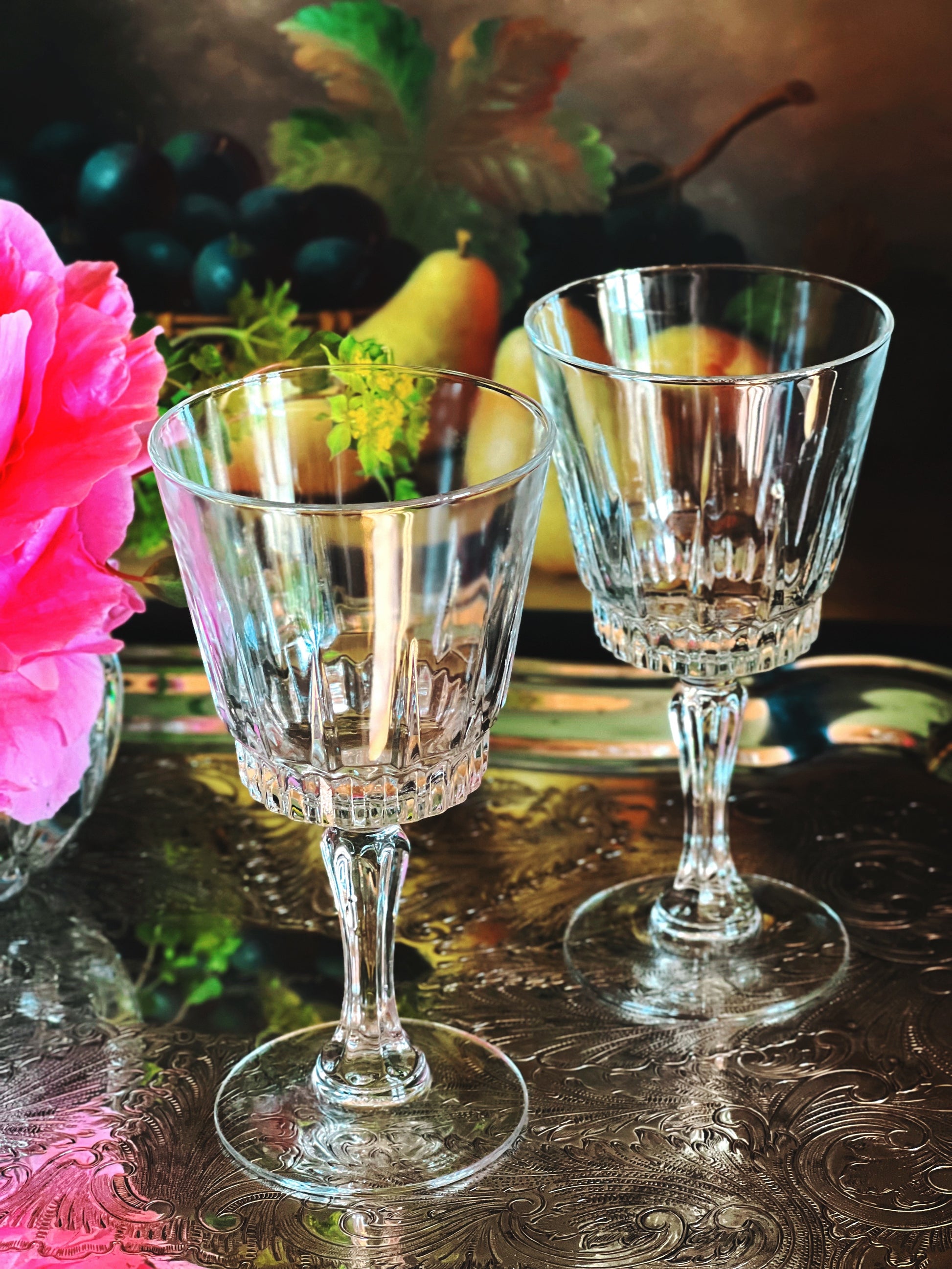 Two crystal glasses on a reflective surface with a floral and fruit background