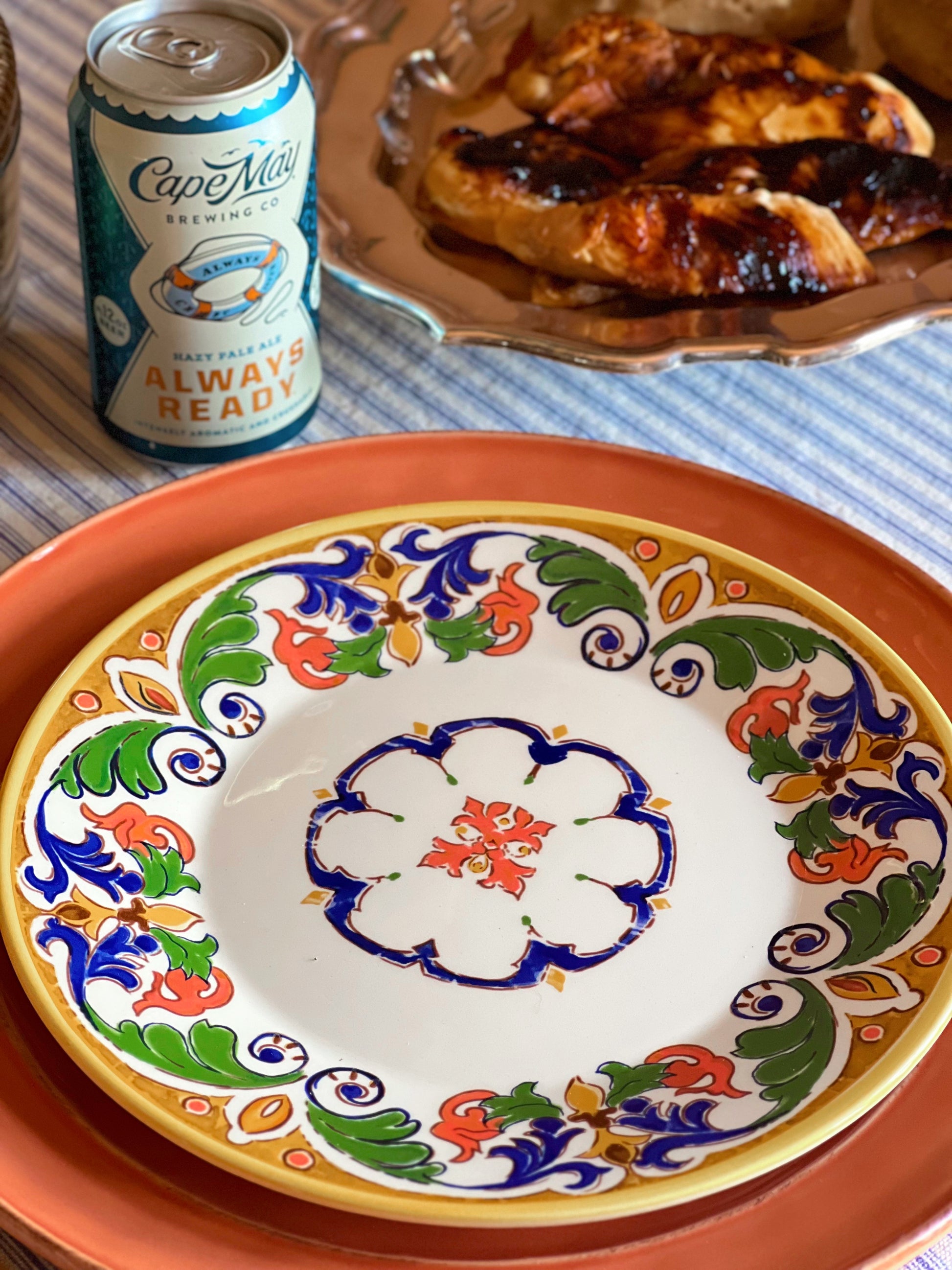 Decorative plate with colorful floral patterns on a table with a can of Cape May beer and grilled chicken.