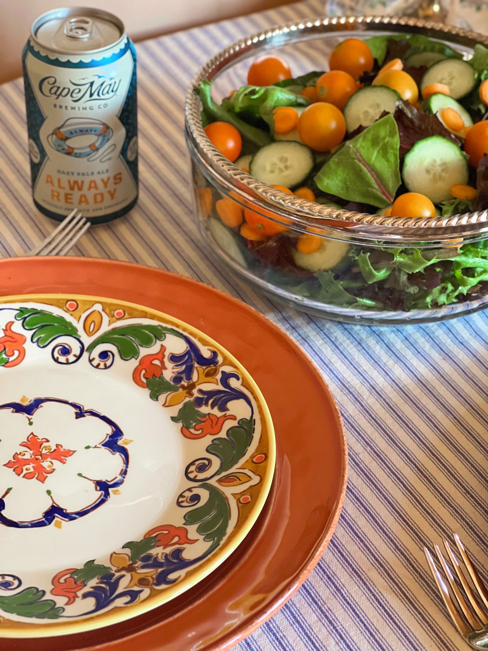 Colorful plates, a can of beer, and a salad on a striped tablecloth.