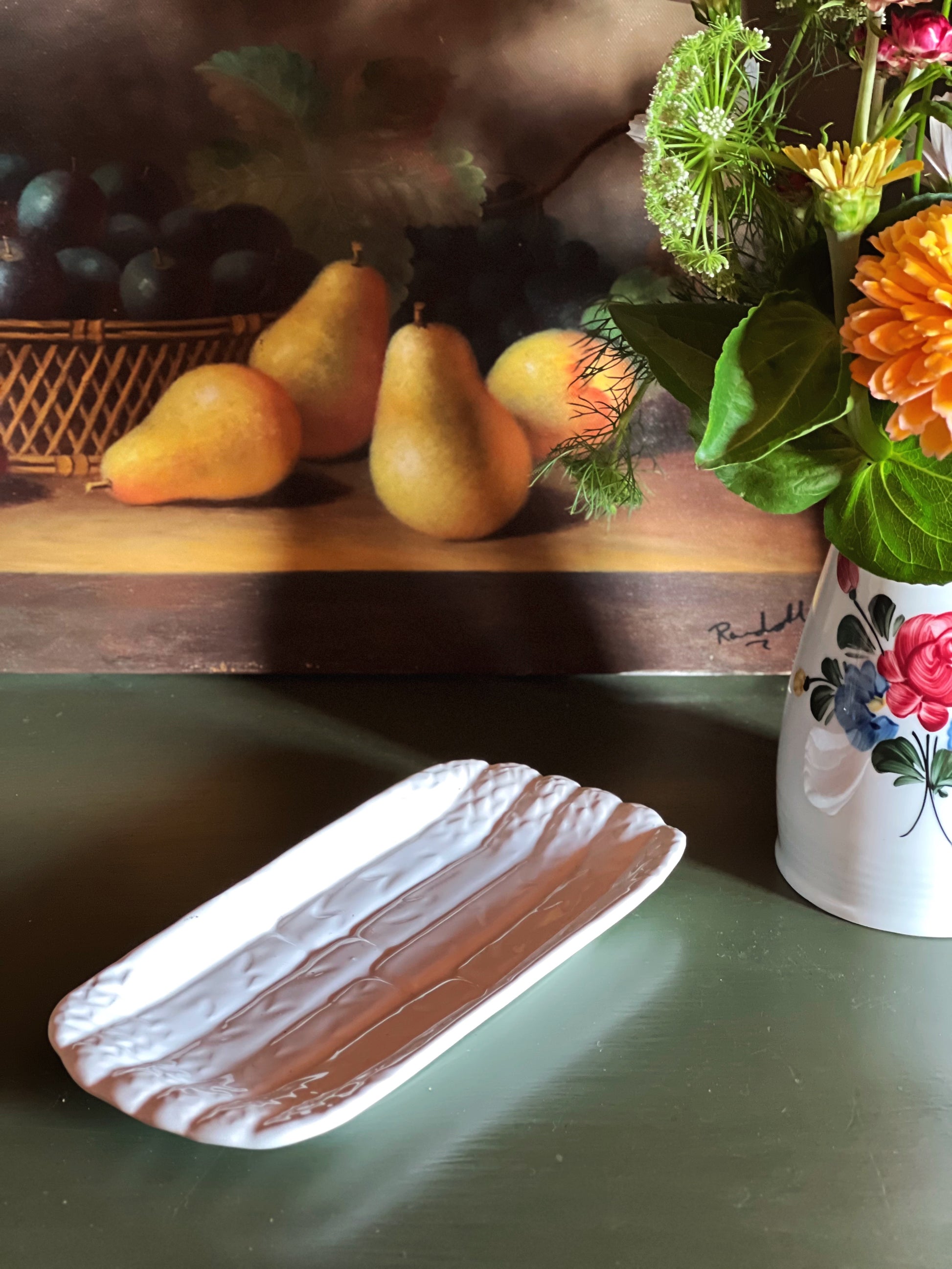 Side view of a vintage Bordallo Pinheiro white majolica asparagus serving dish made in Portugal with sculpted asparagus relief on a green tabletop beside summer flowers.