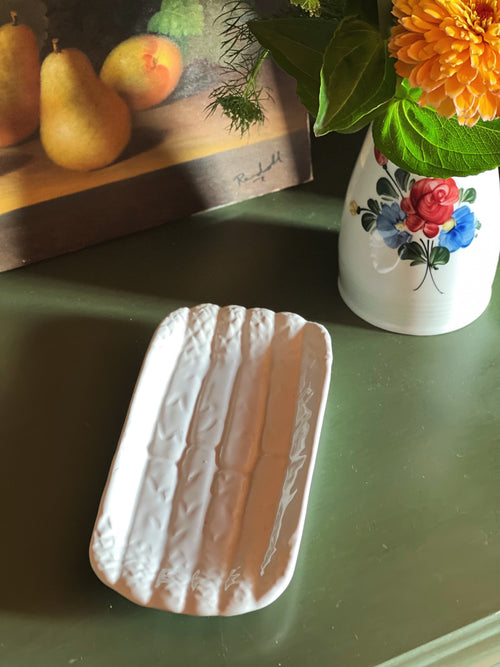 Vintage Bordallo Pinheiro white majolica asparagus serving dish made in Portugal with sculpted asparagus relief on a green tabletop beside summer flowers.