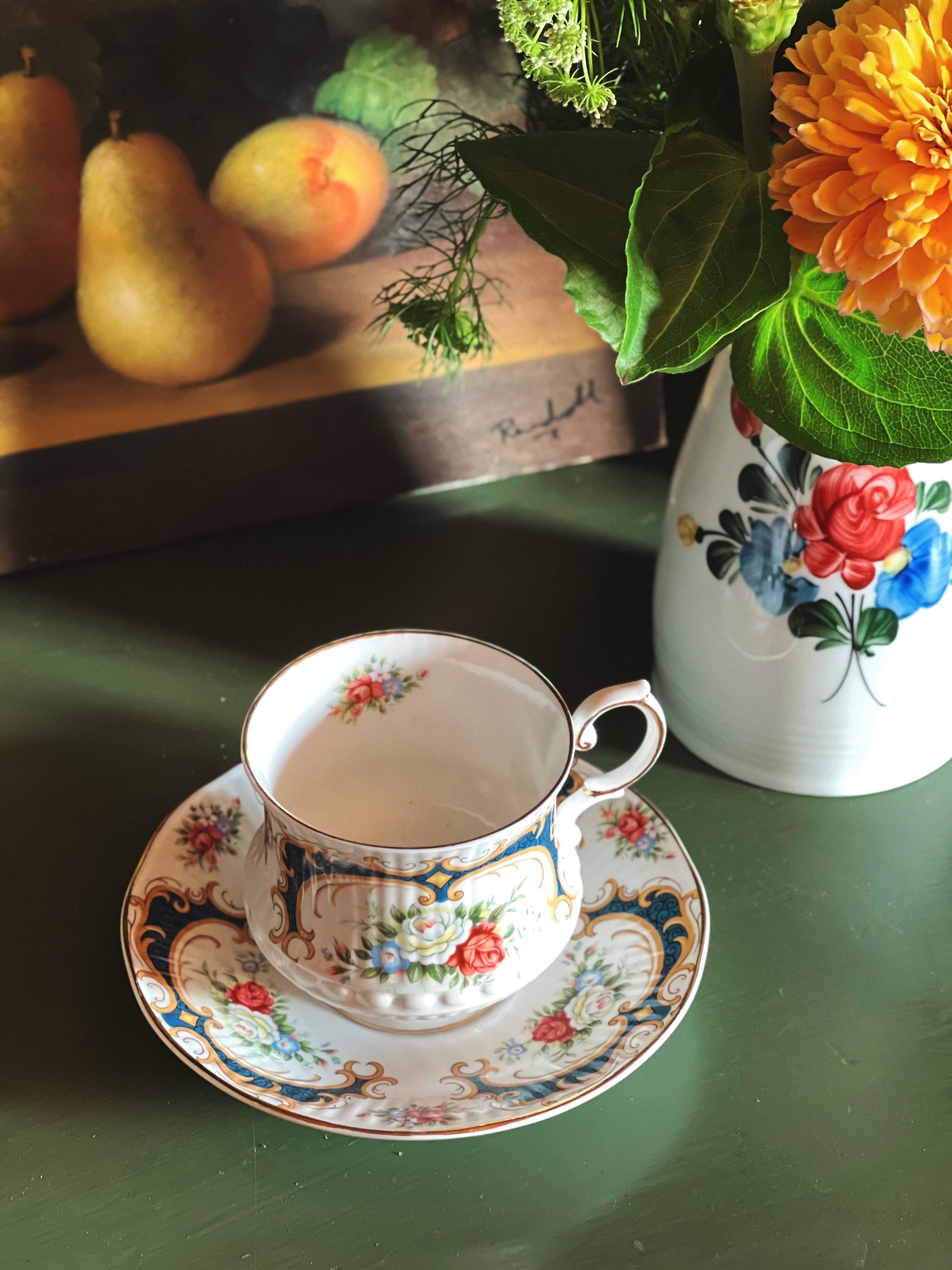 Overhead view of a vintage Queen’s Rosina bone china teacup and saucer with pink floral pattern, navy blue accents, and gold trim styled with fresh flowers and pears on a green tabletop.