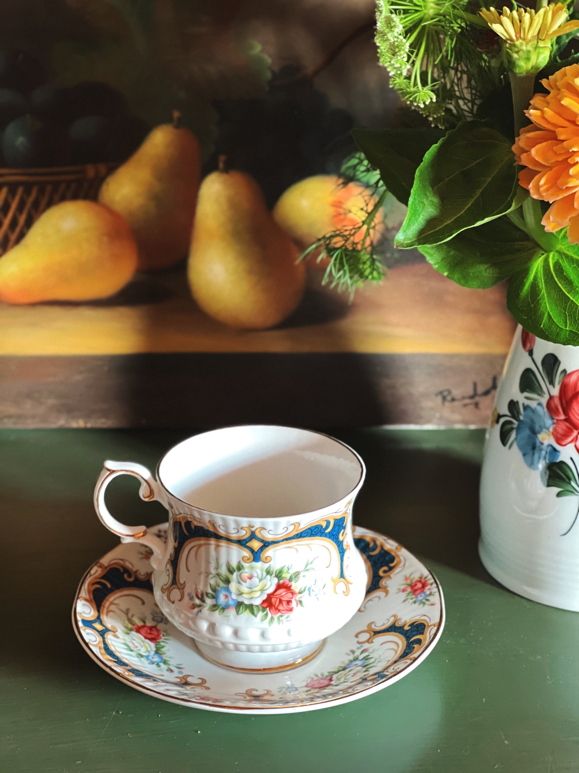 Vintage Queen’s Rosina bone china teacup and saucer with pink floral pattern, navy blue accents, and gold trim styled with fresh flowers and pears on a green tabletop.