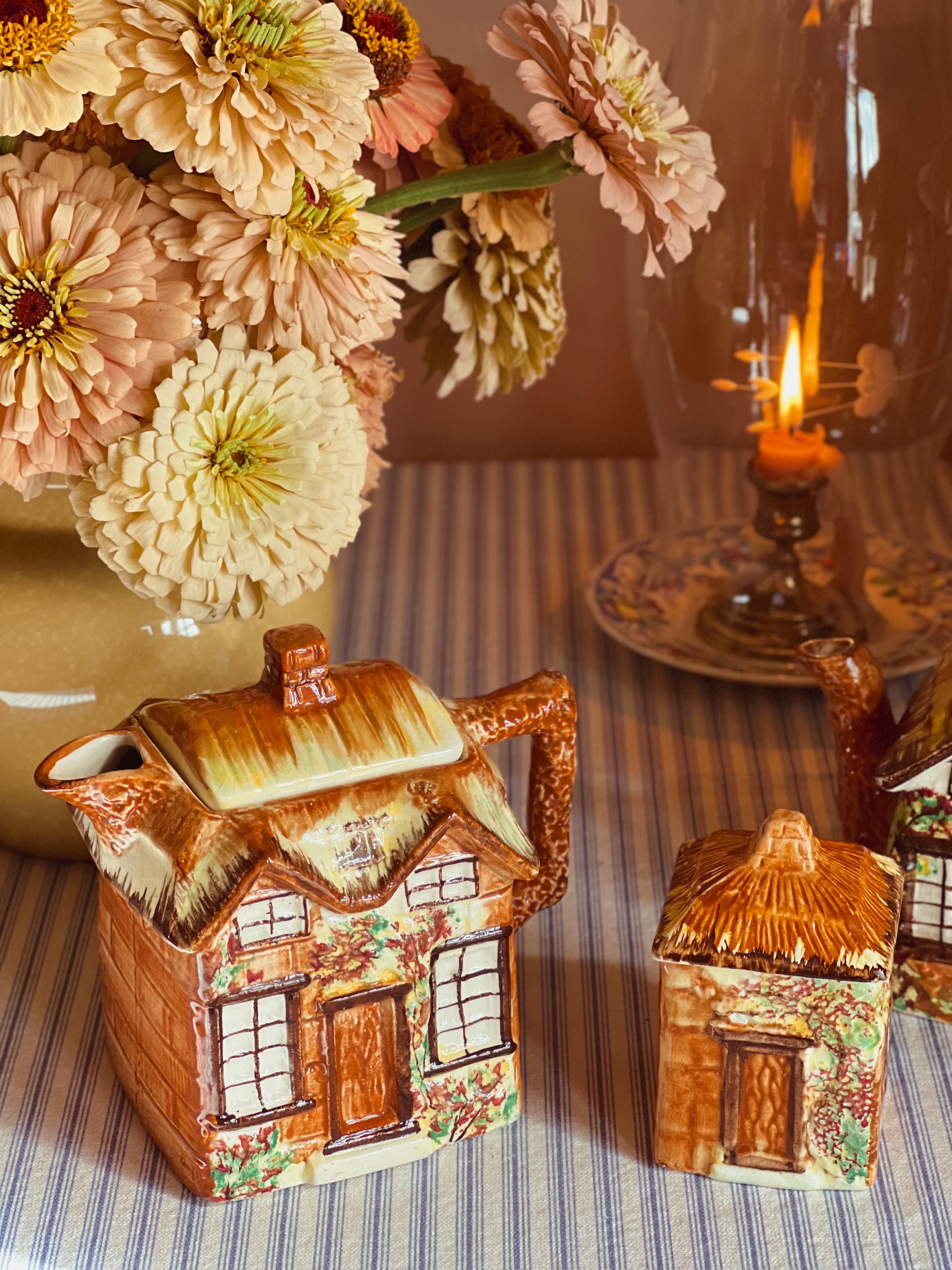 Ceramic house-shaped teapot and container on a table with flowers and candle in the background