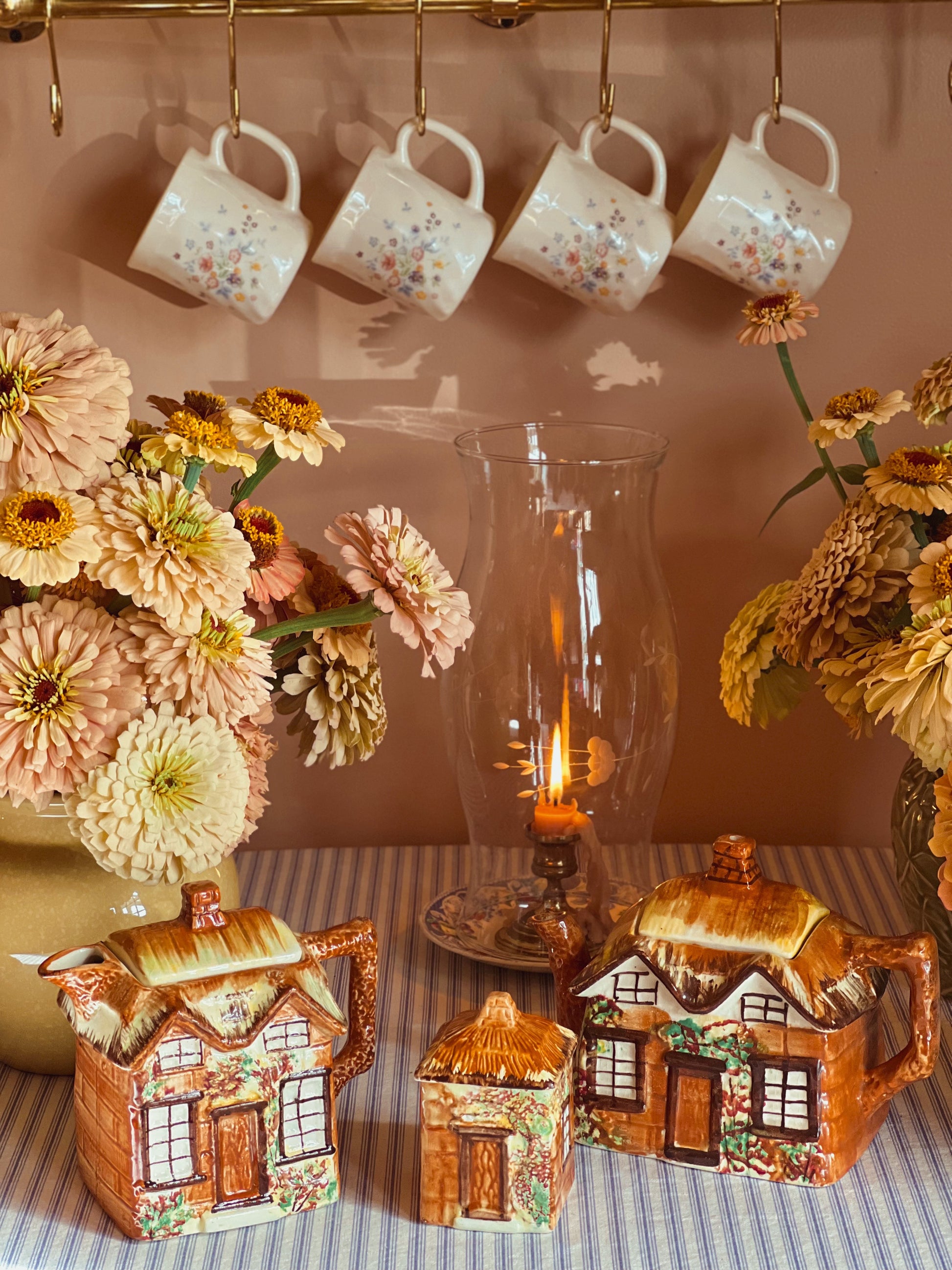 Decorative scene with ceramic house-shaped teapots, mugs hanging on a rack, and floral arrangements.