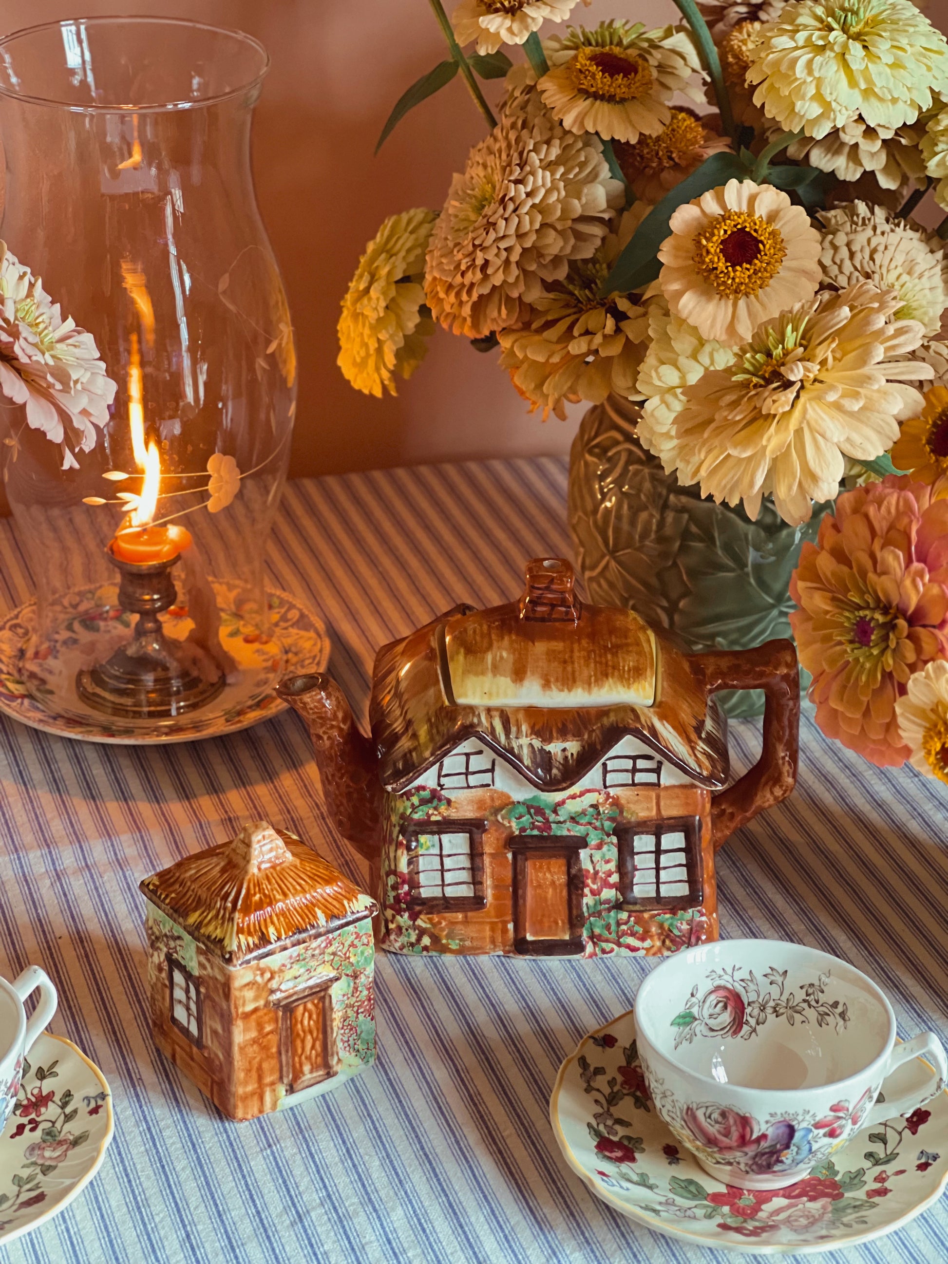 Vintage-style tea set with ceramic house, floral arrangement, and lit lantern on a striped tablecloth.