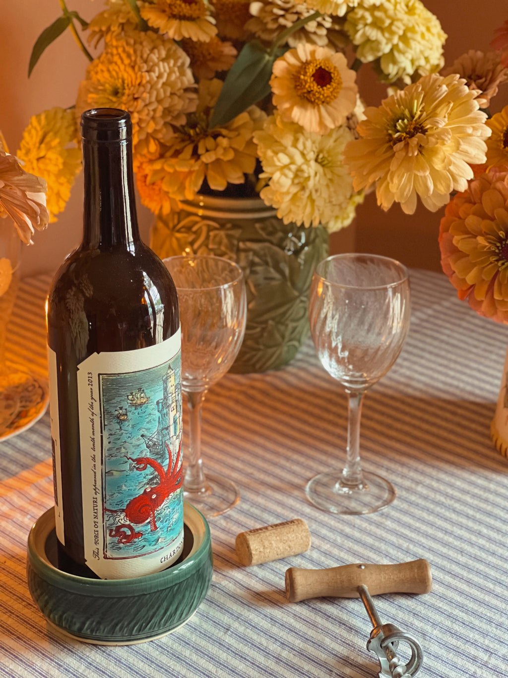 Wine bottle with decorative label on a Michael Gershman Handmade Pottery Wine Coaster , surrounded by flowers and glasses on a table.