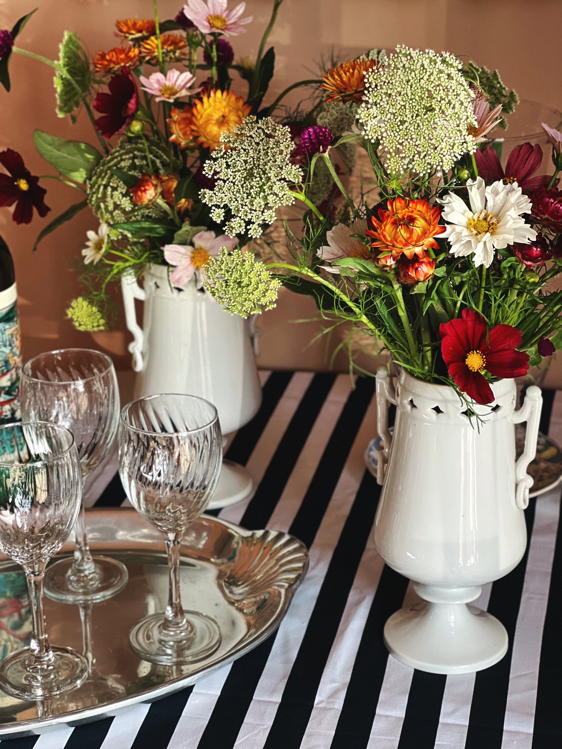 A styled tabletop arrangement with two white ceramic vases holding mixed wildflowers, set on a black-and-white striped tablecloth with clear wine glasses and a silver tray.