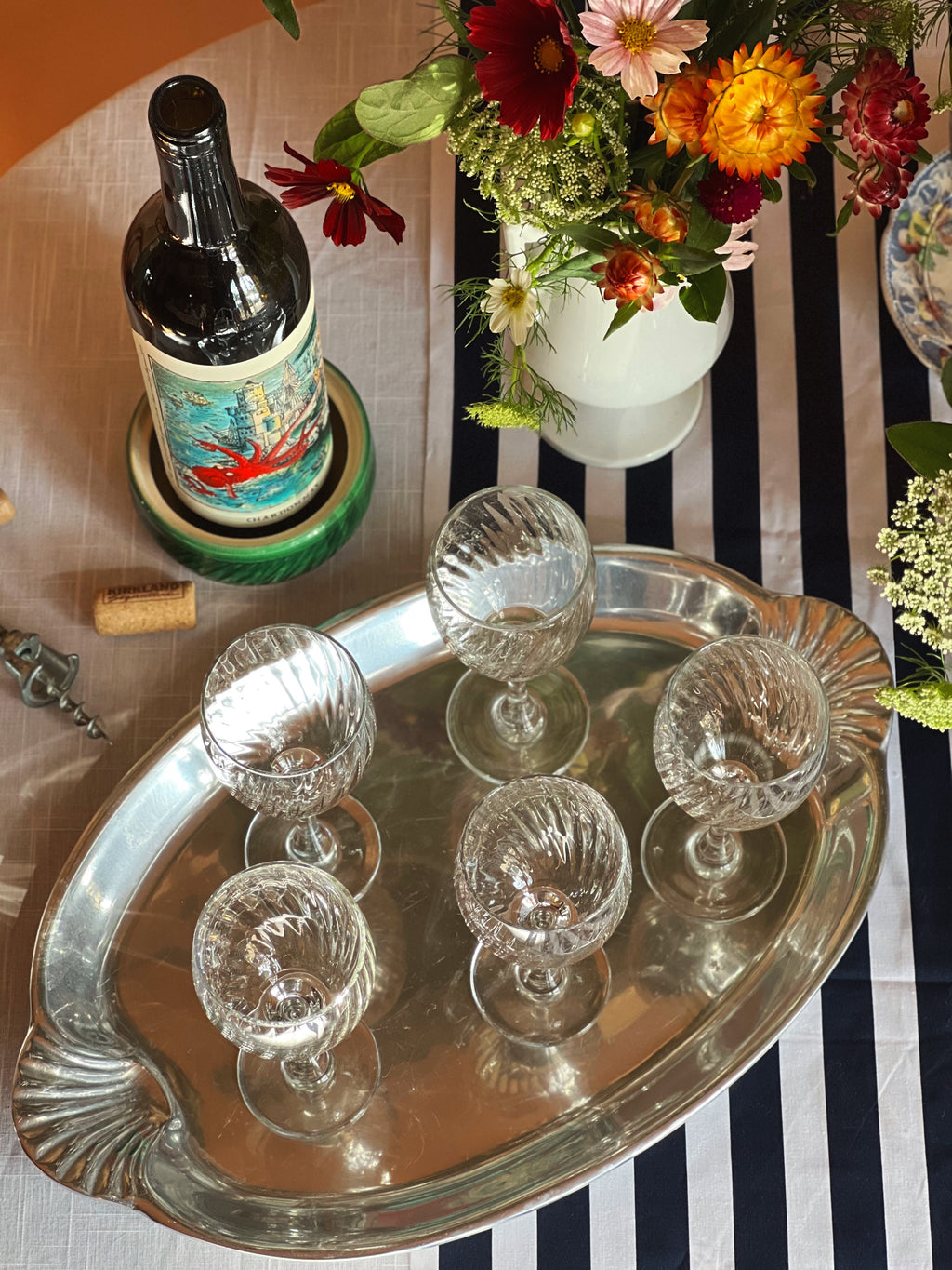 Wilton Armetale oval tray with scallop shell handles with crystal glasses, a bottle of wine, and a floral arrangement on a striped tablecloth.