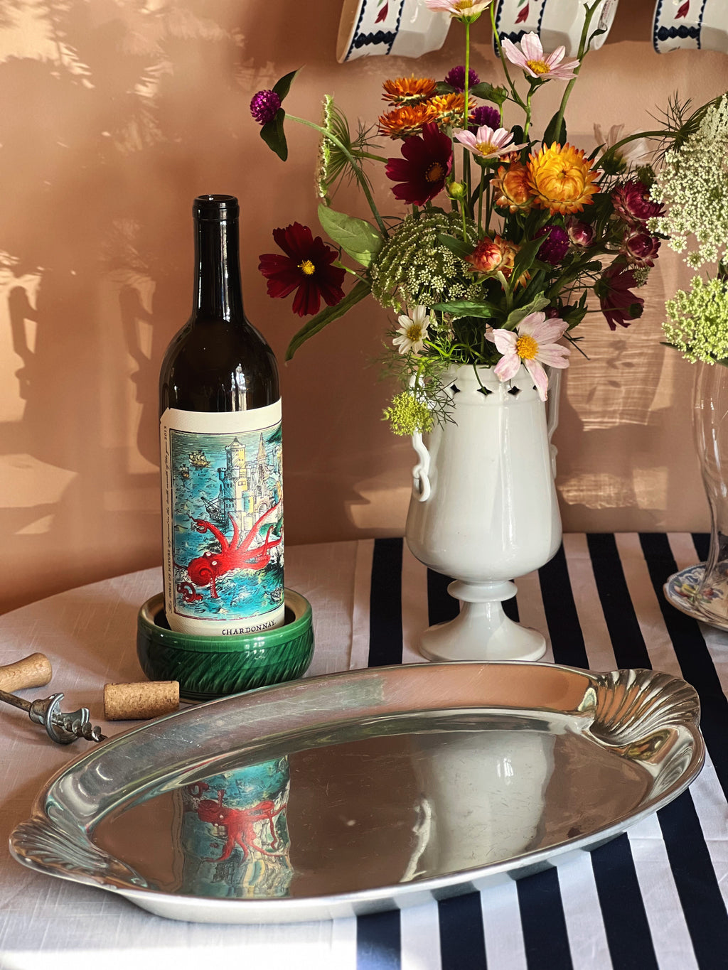 Wine bottle with decorative label, Wilton Armetale platter with scallop shell handles, and floral arrangement on a striped tablecloth.