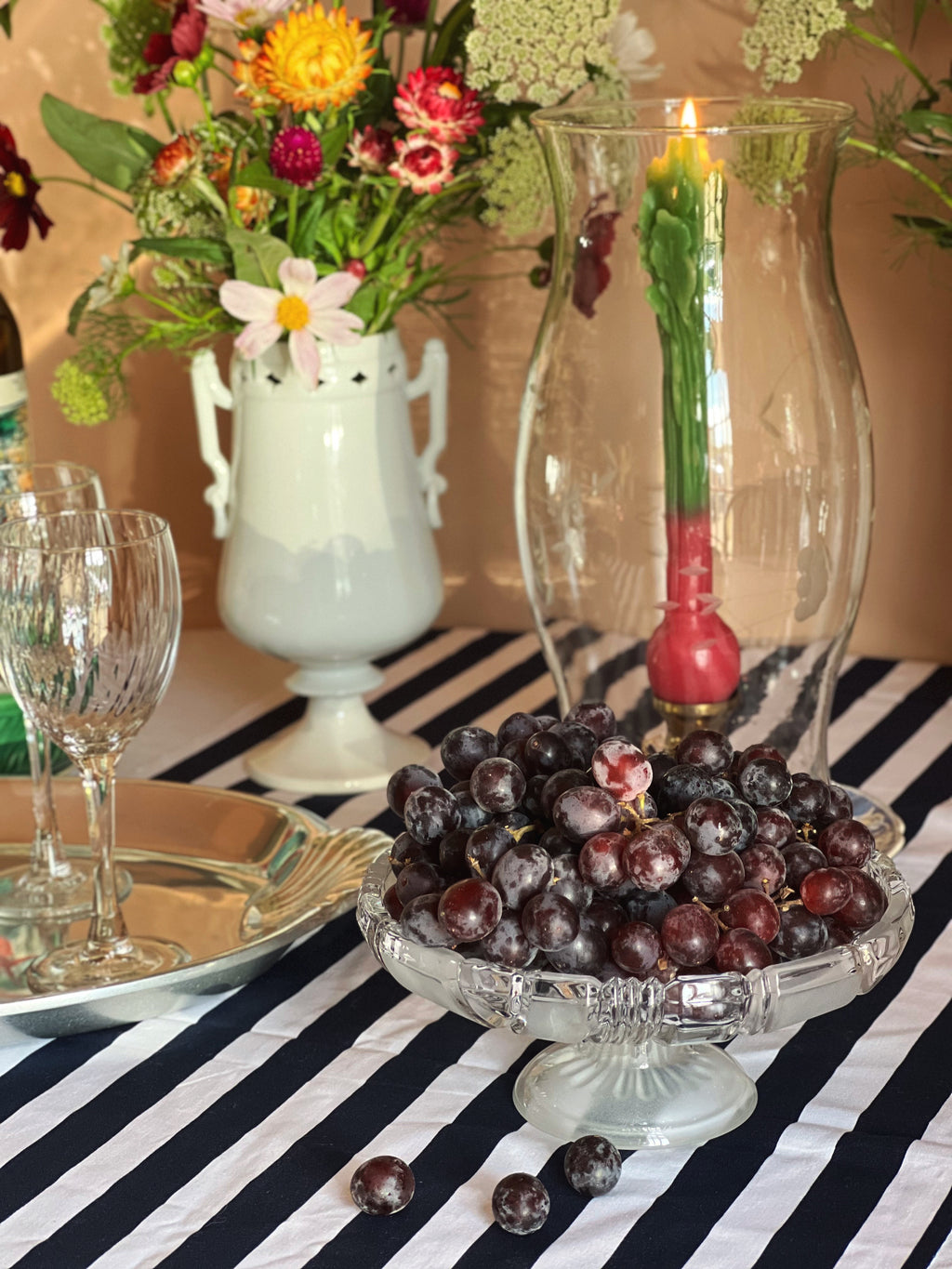 A styled tabletop scene with a clear glass pedestal bowl filled with dark purple grapes in the foreground, a tall clear glass vase with a lit candle, a white ceramic pitcher of flowers, and a black-and-white striped tablecloth.